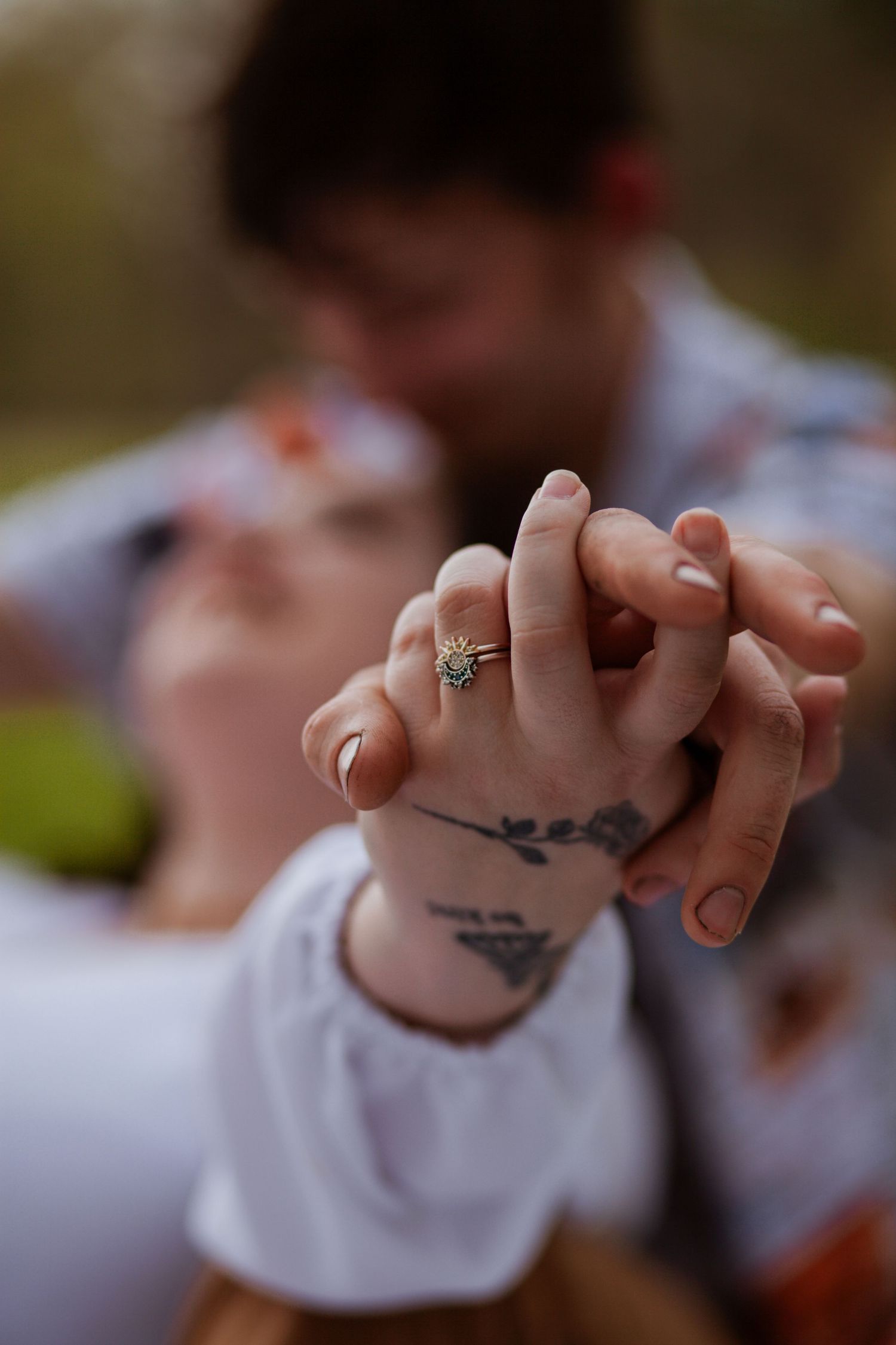 Close-up of intertwined hands with engagement ring and tattoo visible.