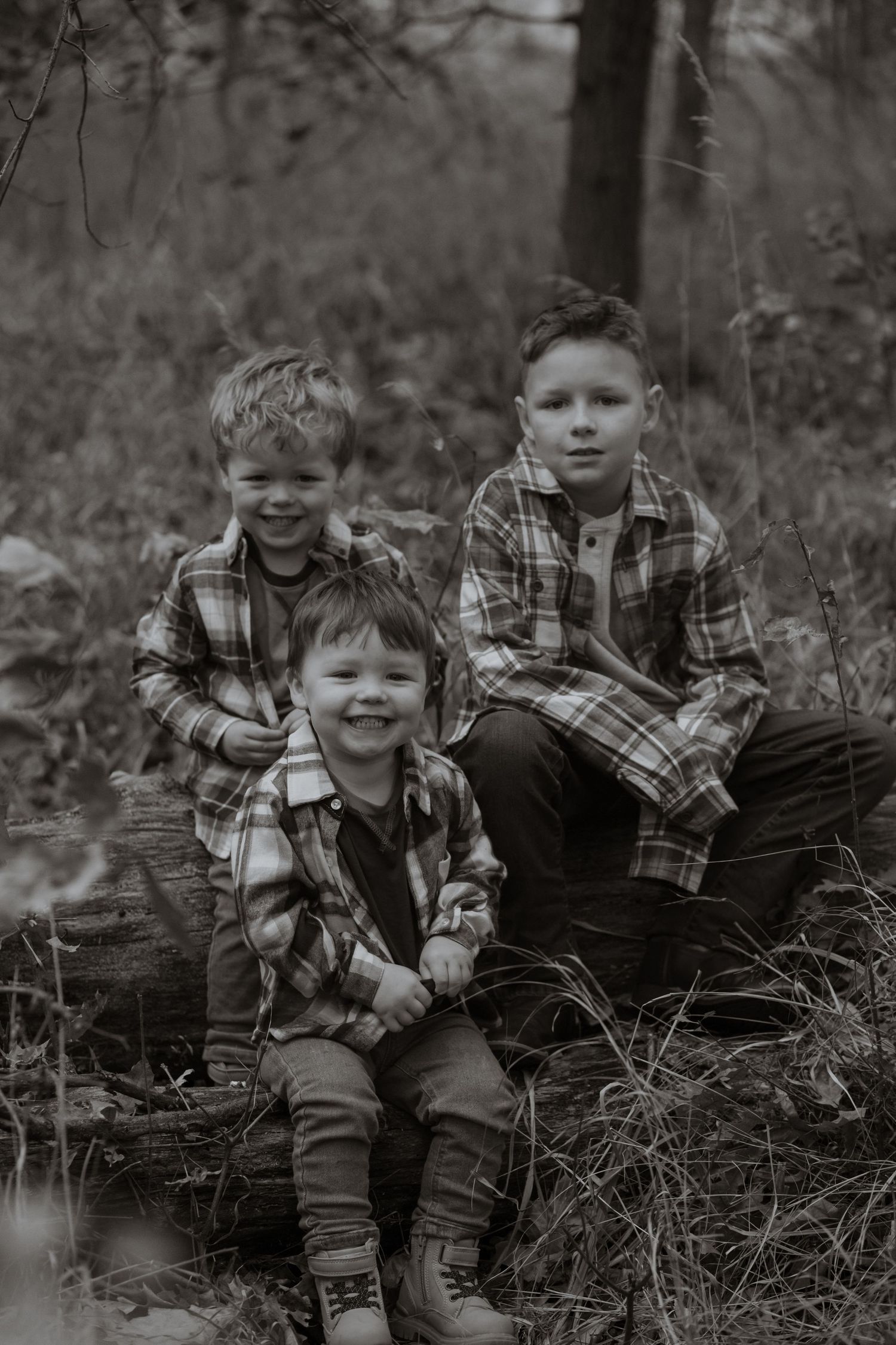 Two children in plaid shirts sit together on a fallen log in a black and white forest scene.