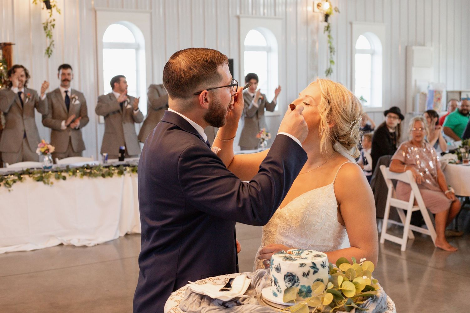 A wedding reception moment during cake cutting in a white-walled venue with guests watching.