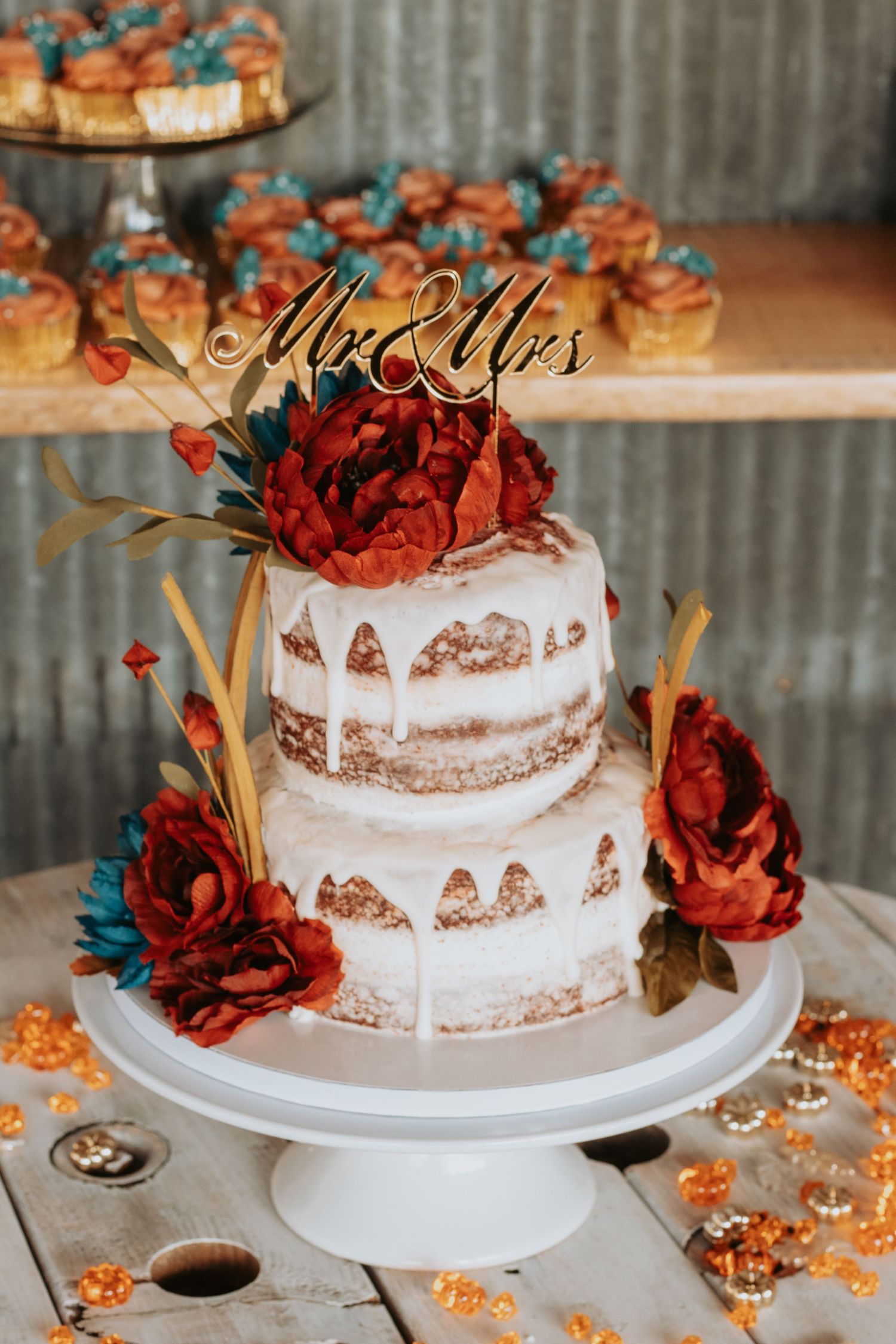 Rustic wedding cake decorated with red and teal flowers on white stand.