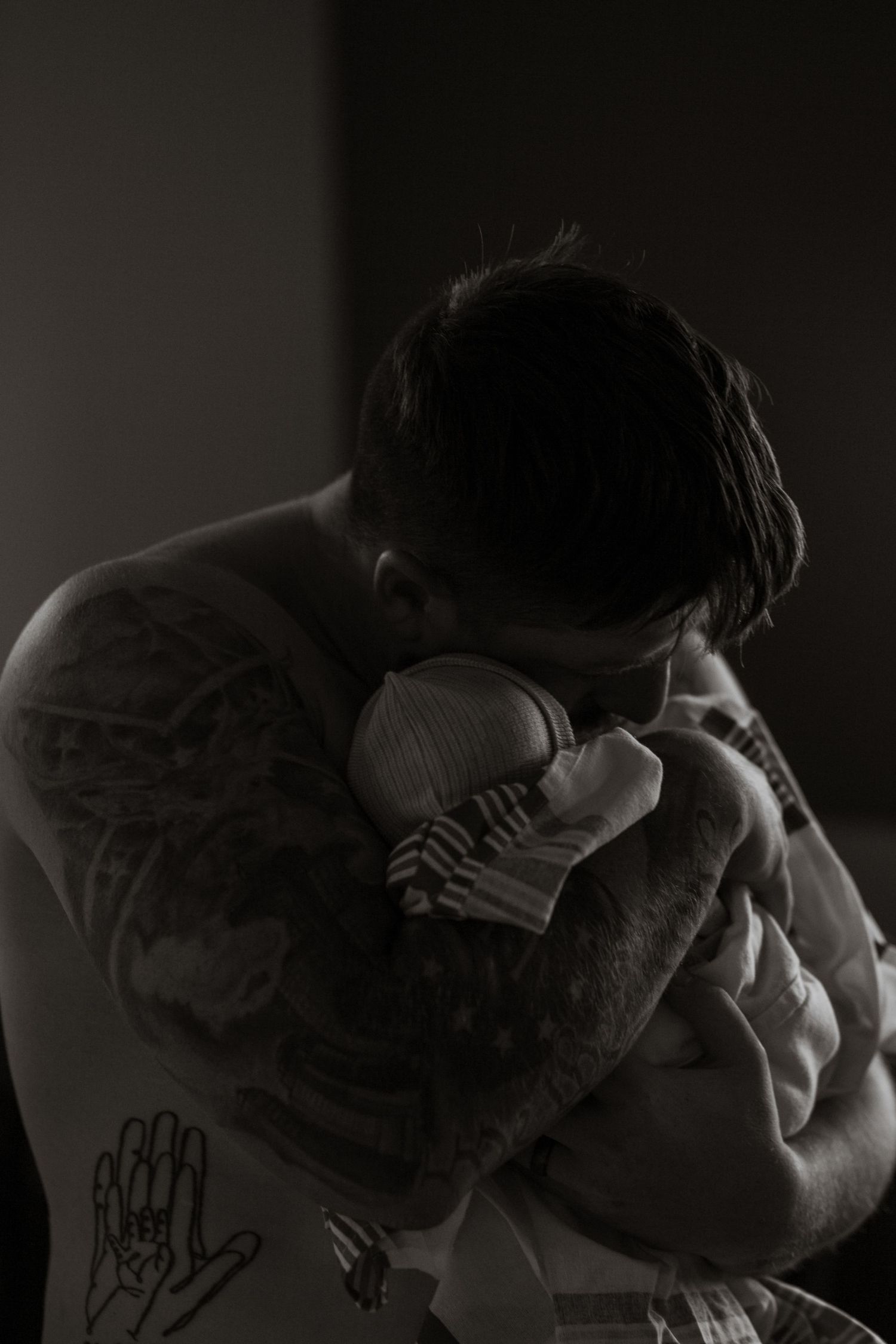 Moody black and white photo of a tattooed person holding a newborn baby in dark lighting.