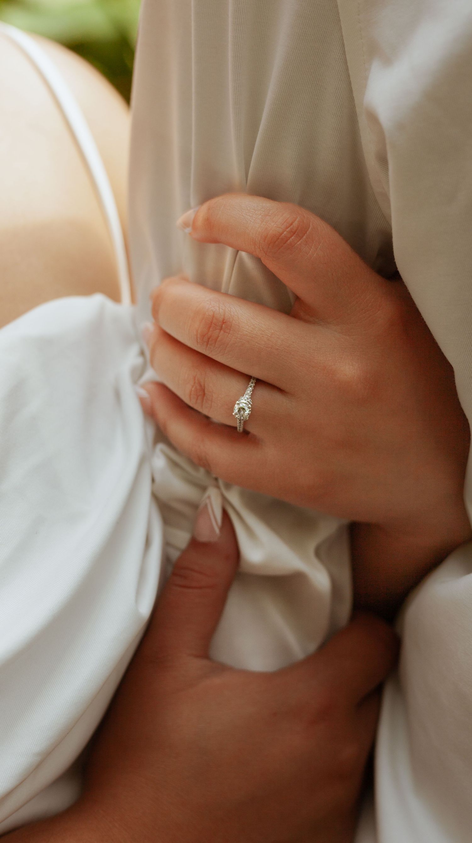 Close-up of engagement ring and hands against white fabric in soft lighting.