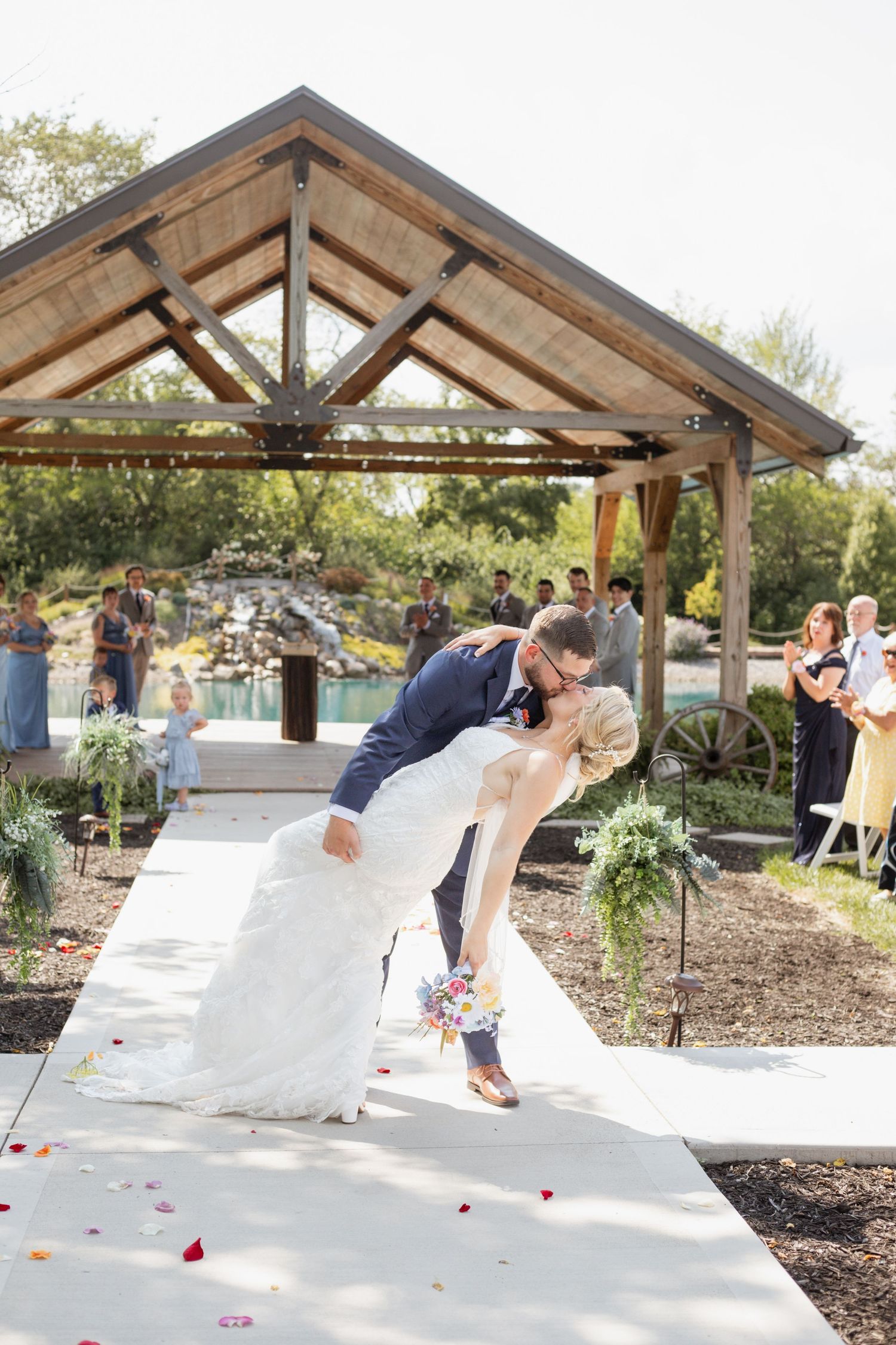 Dramatic wedding dip kiss under wooden pavilion with guests watching in background.
