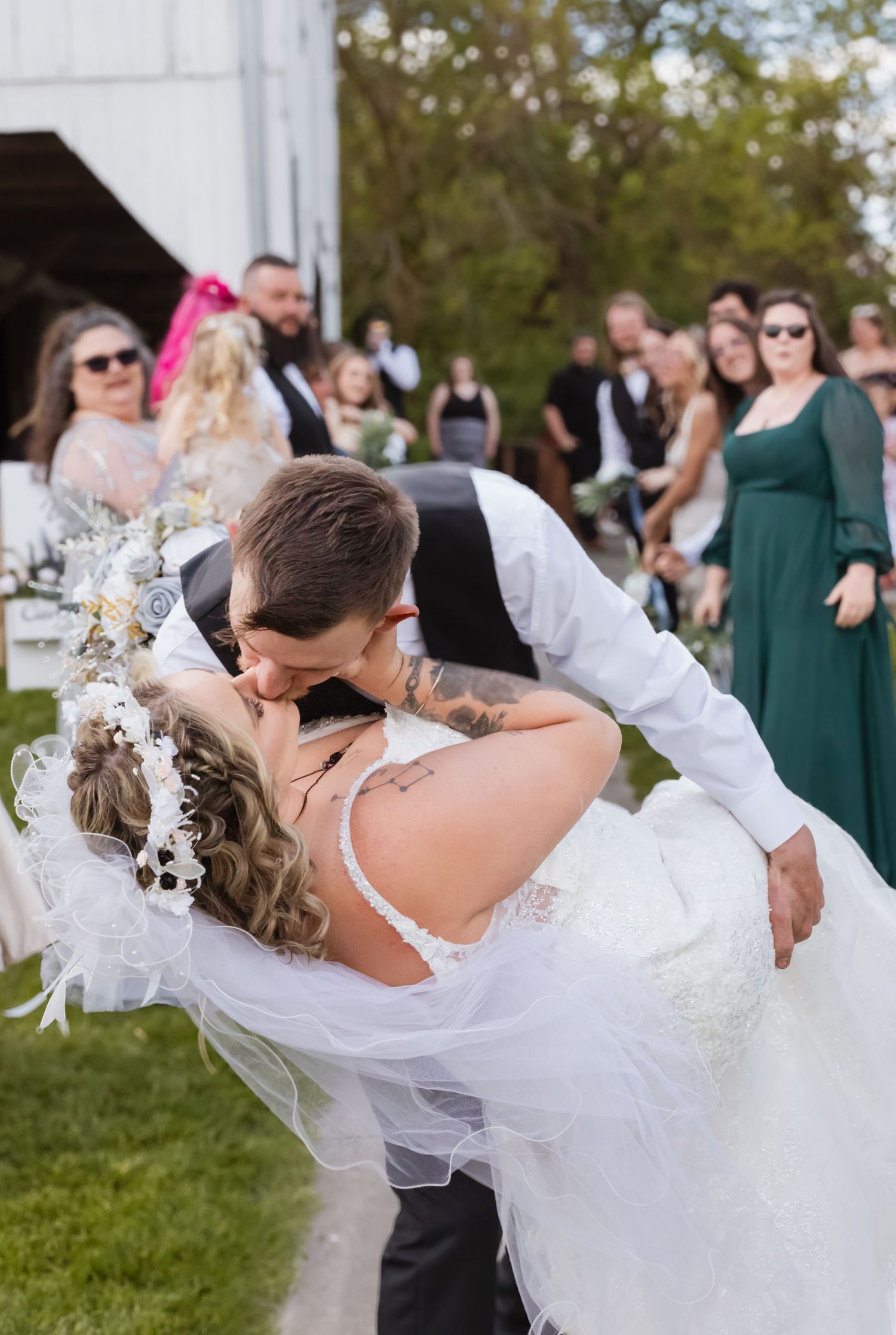 A romantic dip kiss between newlyweds surrounded by wedding guests on a sunny day.