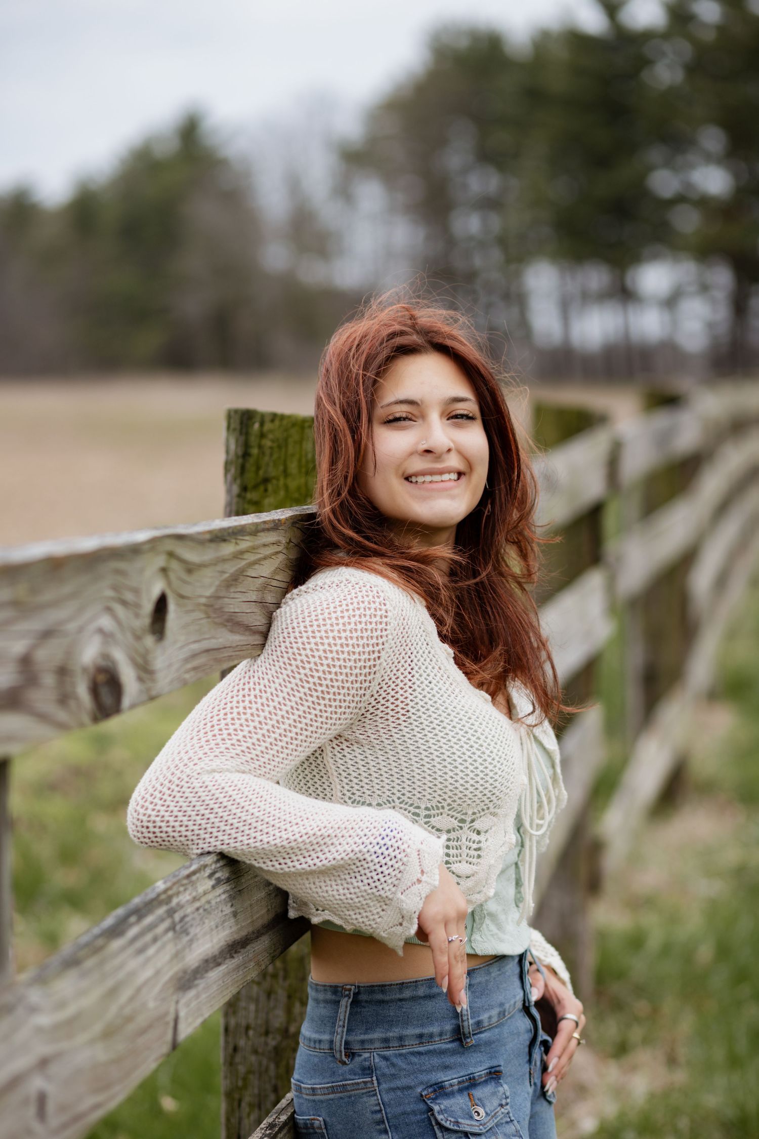 Person in white knit sweater and jeans poses playfully against wooden fence in outdoor setting.