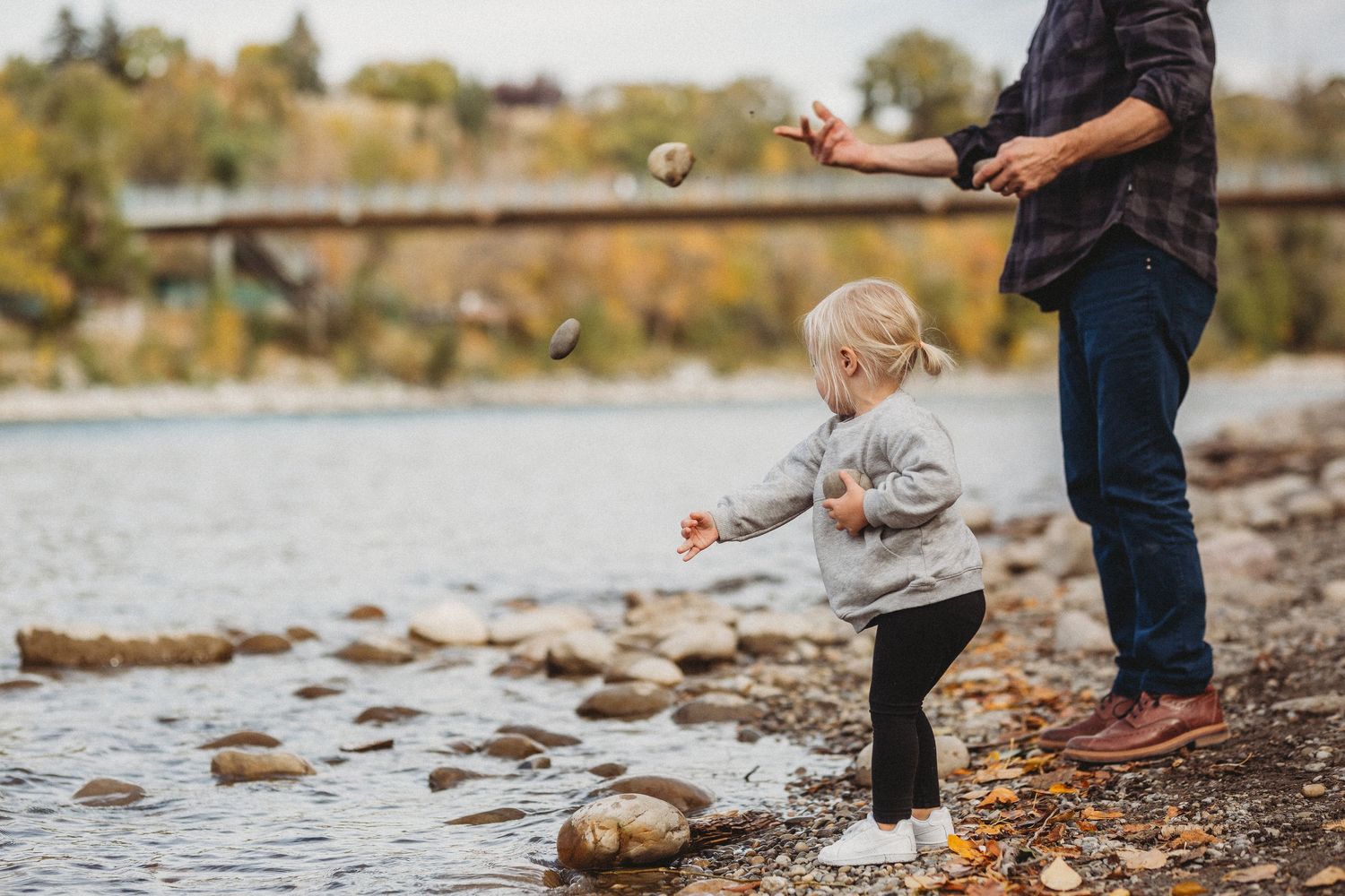 Toddler girl and grandfather throwing rocks in the river in a documentary style Calgary family photography session in Prince's Island Park