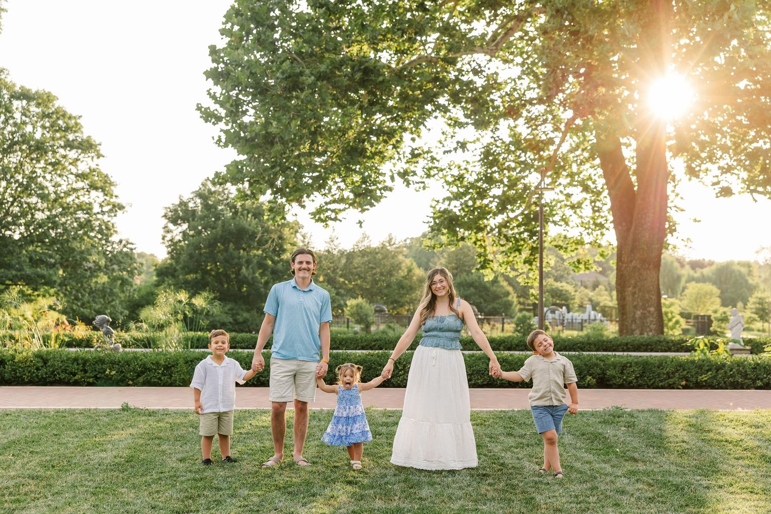 Family of five holding hands on green lawn at golden hour with sunlit trees in background.