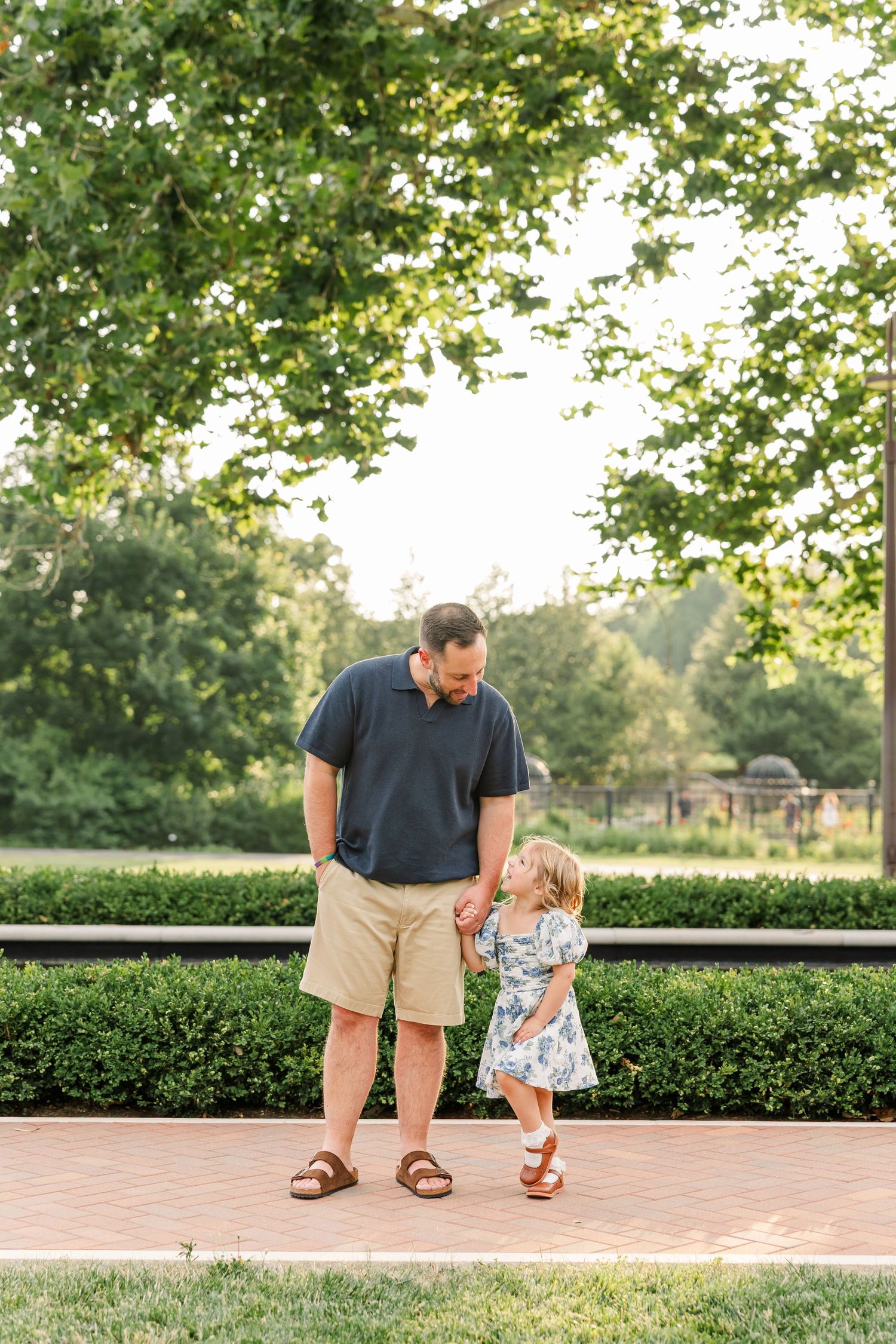 Father and toddler daughter share sweet moment on brick path surrounded by manicured hedges in park.