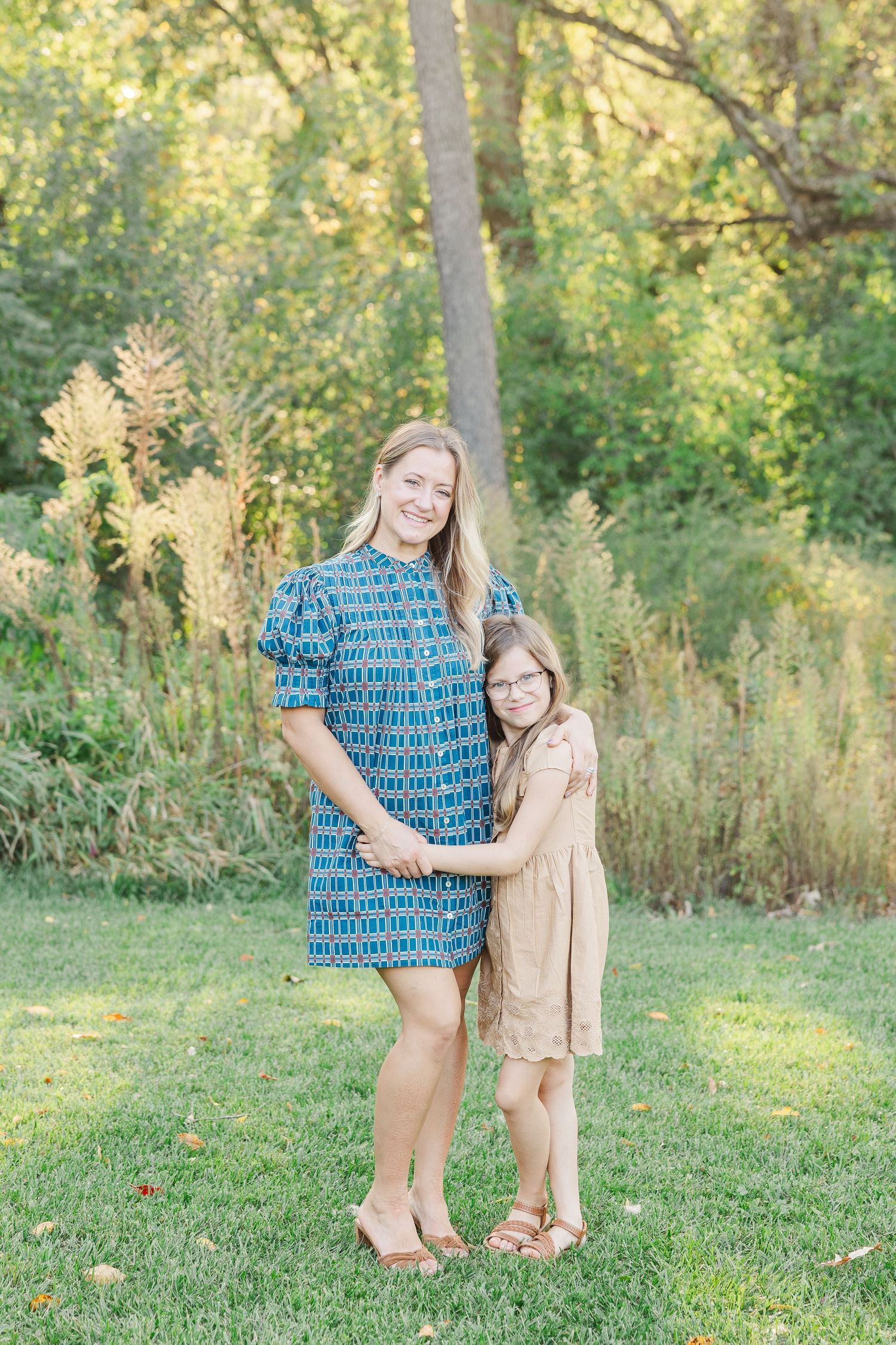 Two people embrace while standing on green grass in a sunlit wooded area during golden hour.