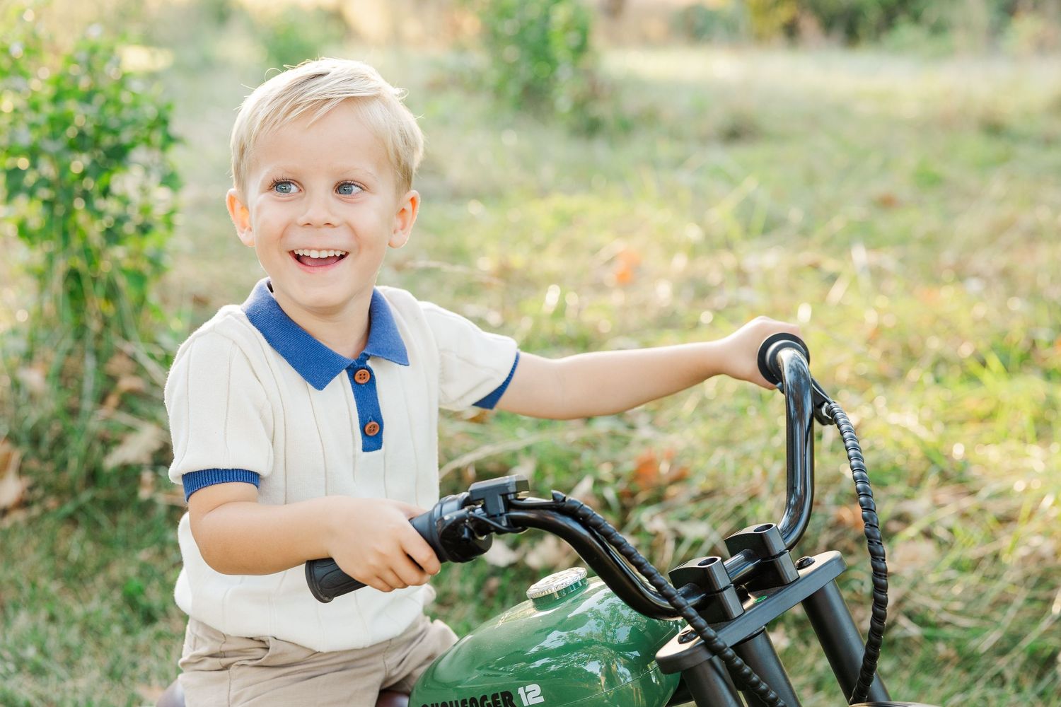 Young child sits on a green toy tractor in a sunny outdoor setting with natural grass and foliage in background.