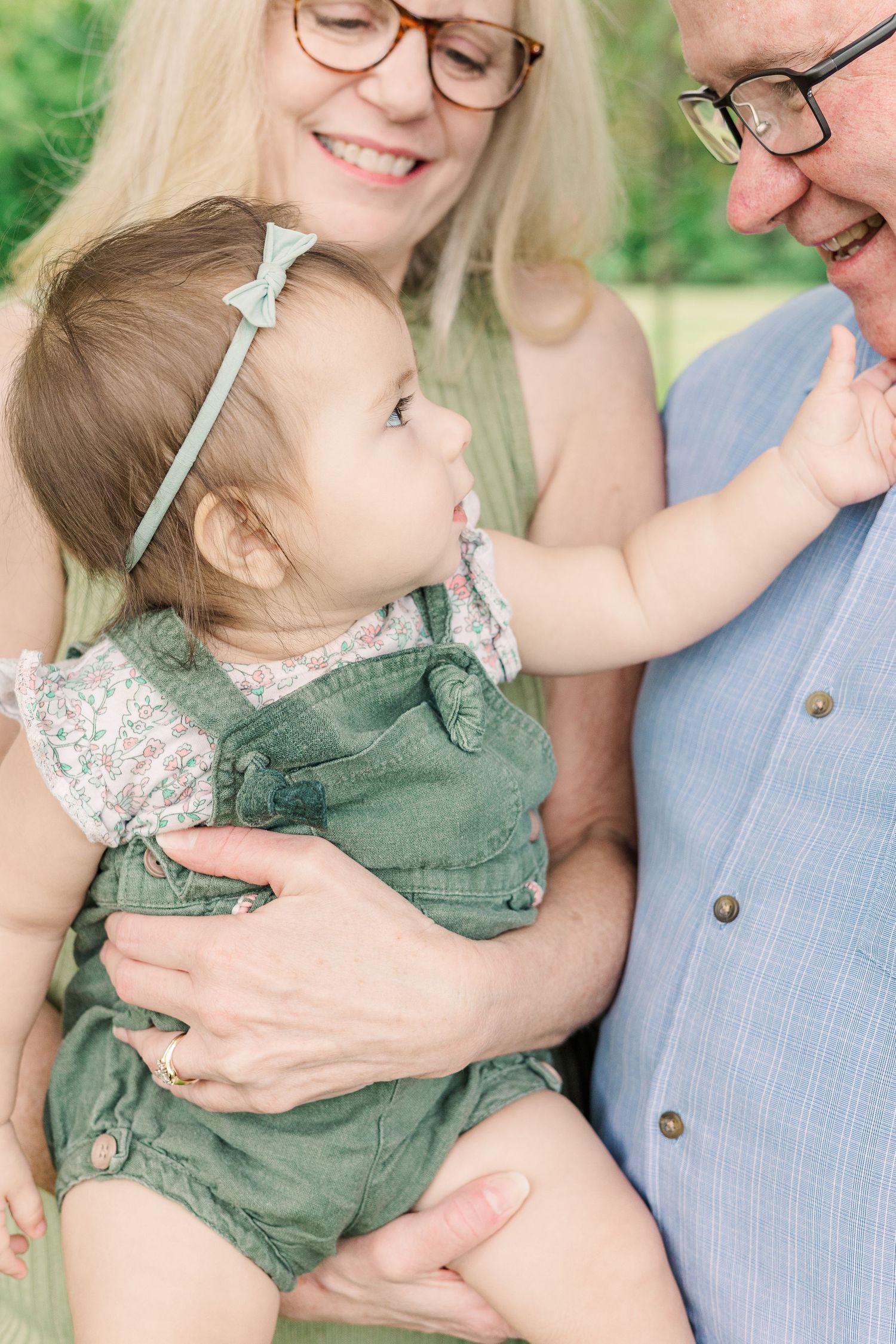 A joyful family moment with a toddler in green overalls being held by adults in a garden setting.