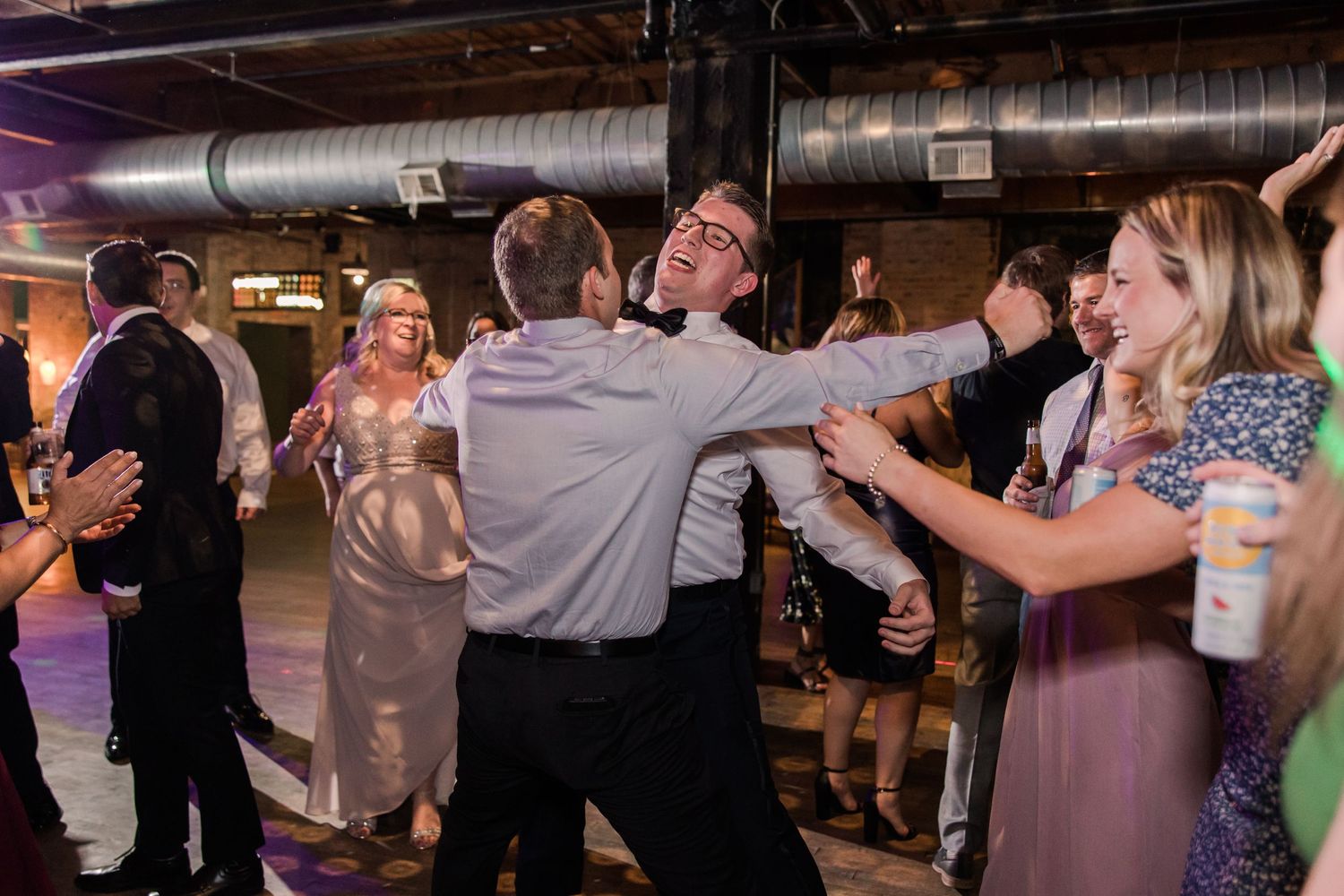 Wedding guests dancing energetically in an industrial-style venue with exposed ductwork and party lighting.