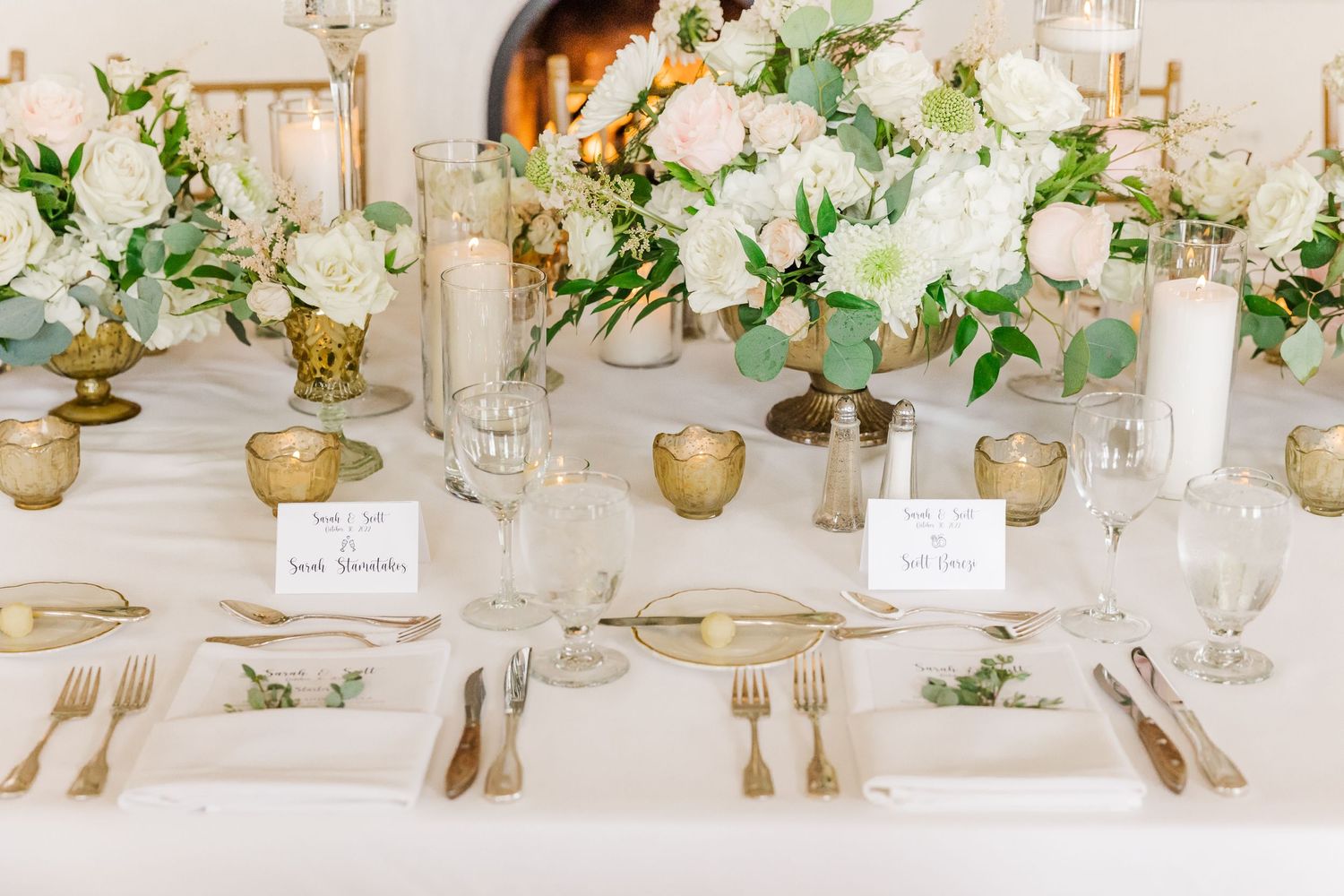 Elegant wedding table setting with white floral centerpieces, gold accents, and crystal glassware on white linens.