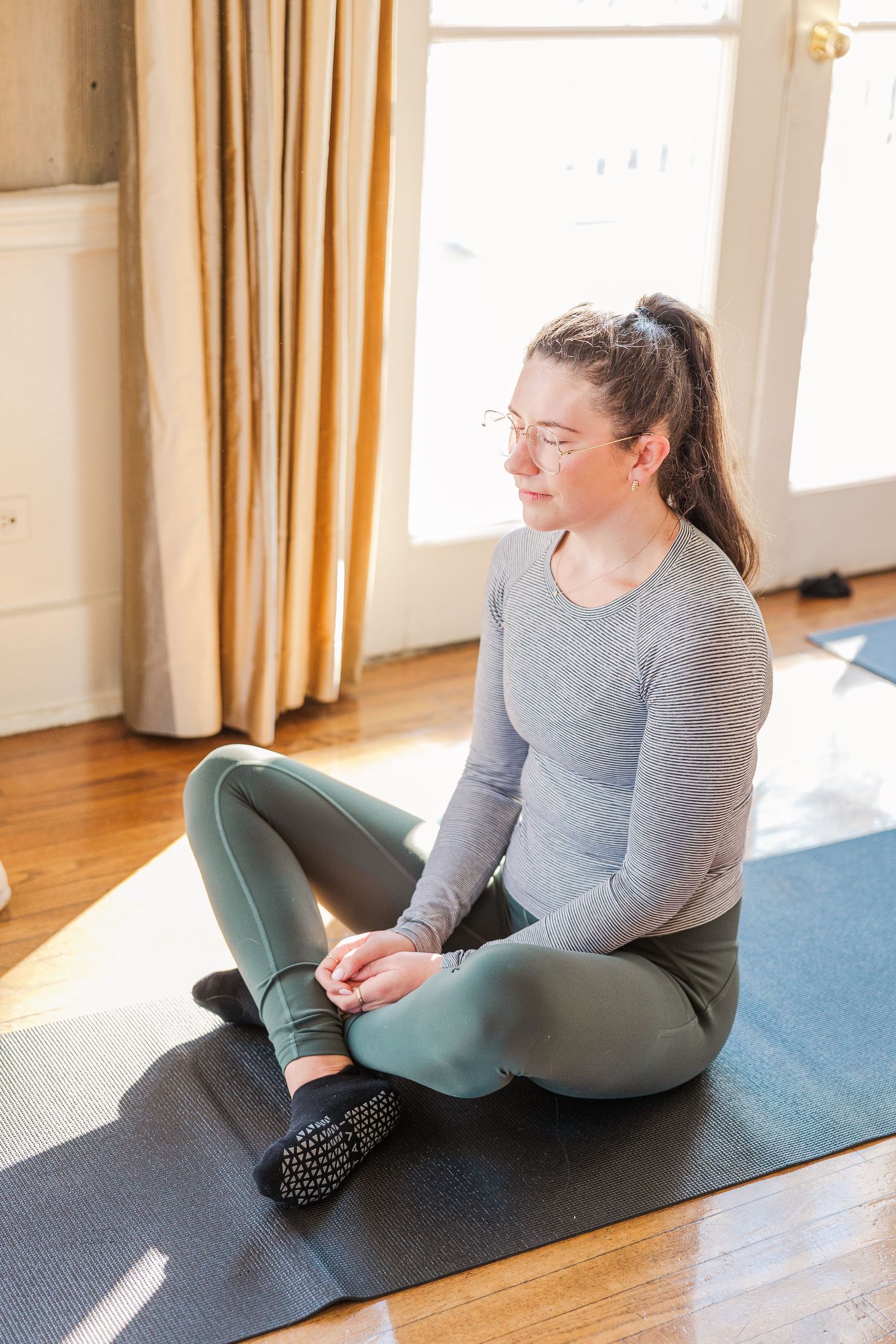 Person sitting cross-legged on a yoga mat in a sunlit room with hardwood floors.