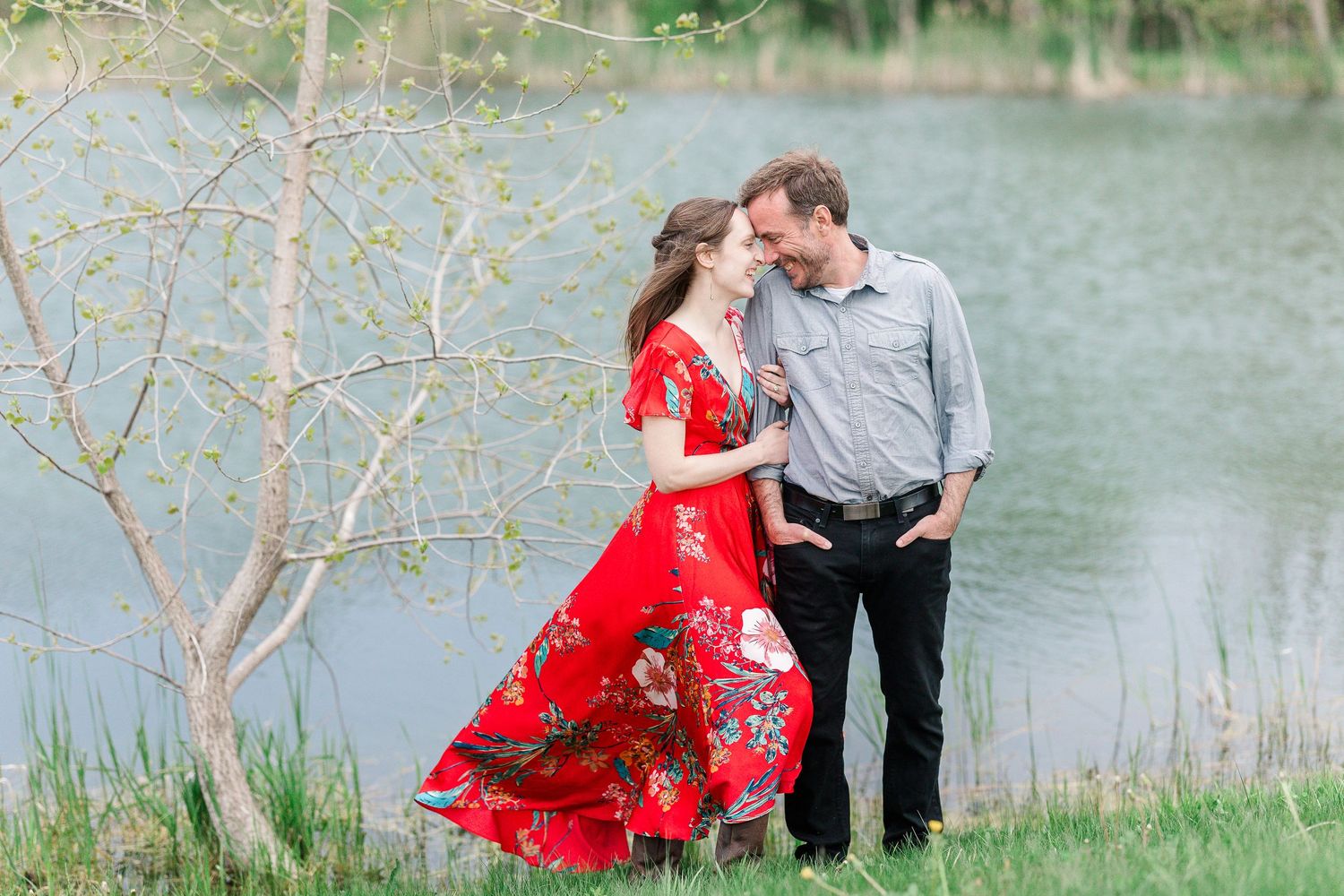 A couple in coordinated outfits stands by a serene lake with spring trees in the background.