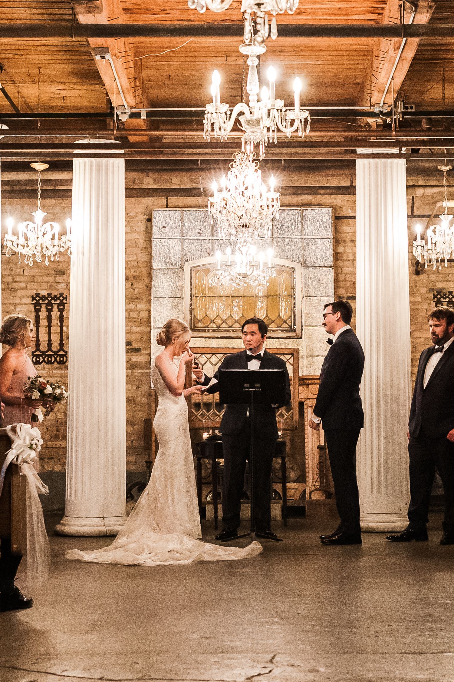 Wedding ceremony in rustic venue with elegant chandeliers and white columns.
