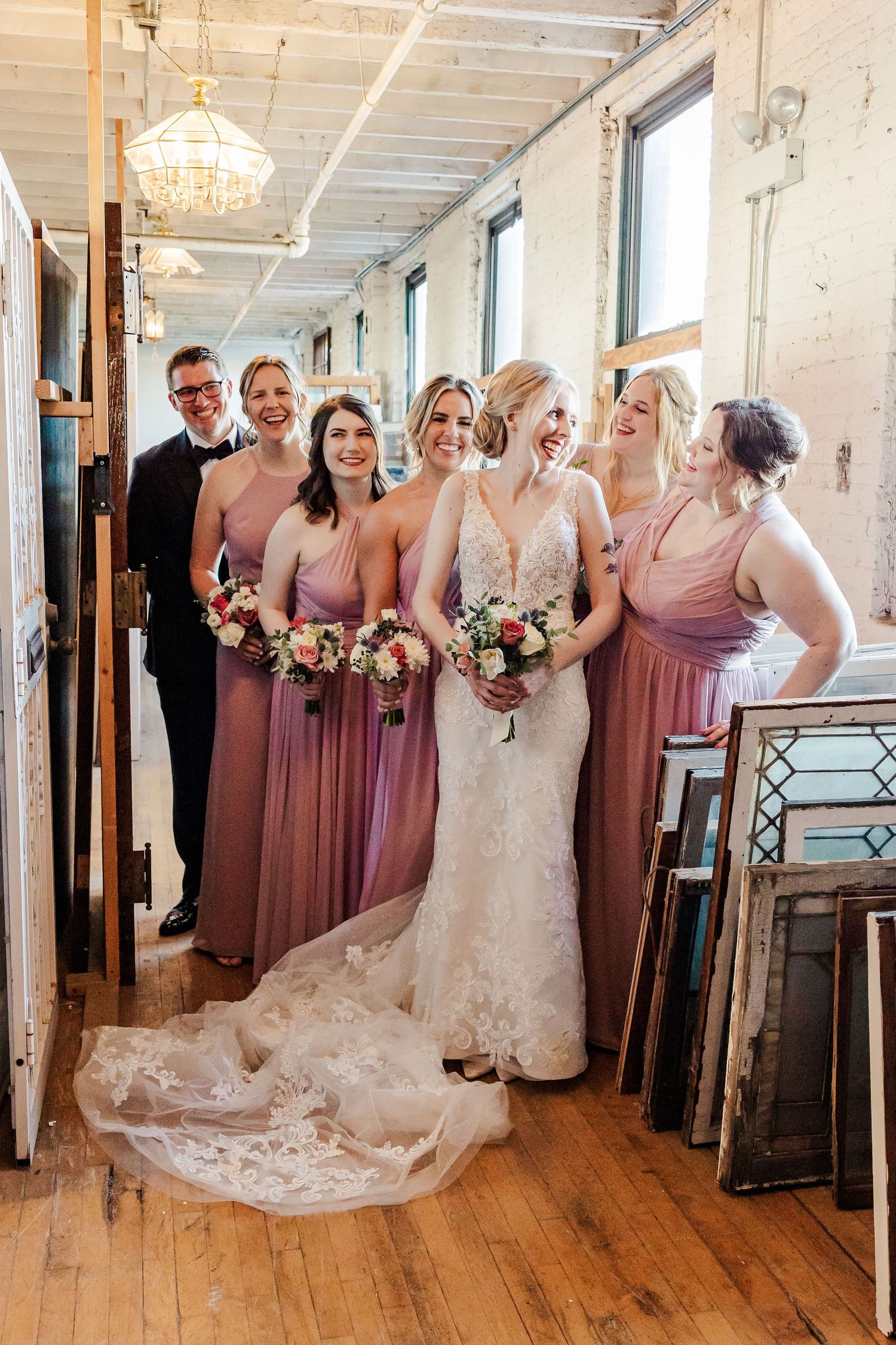 Wedding party poses in rustic hallway with bride in lace gown and bridesmaids wearing dusty rose dresses.