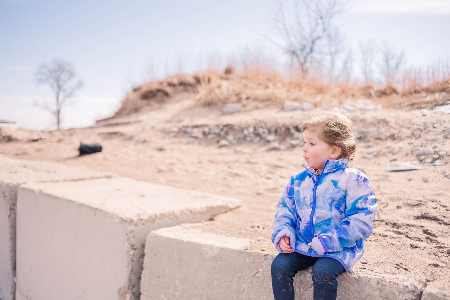 A figure in a blue tie-dye jacket sits on concrete blocks against a sandy landscape.