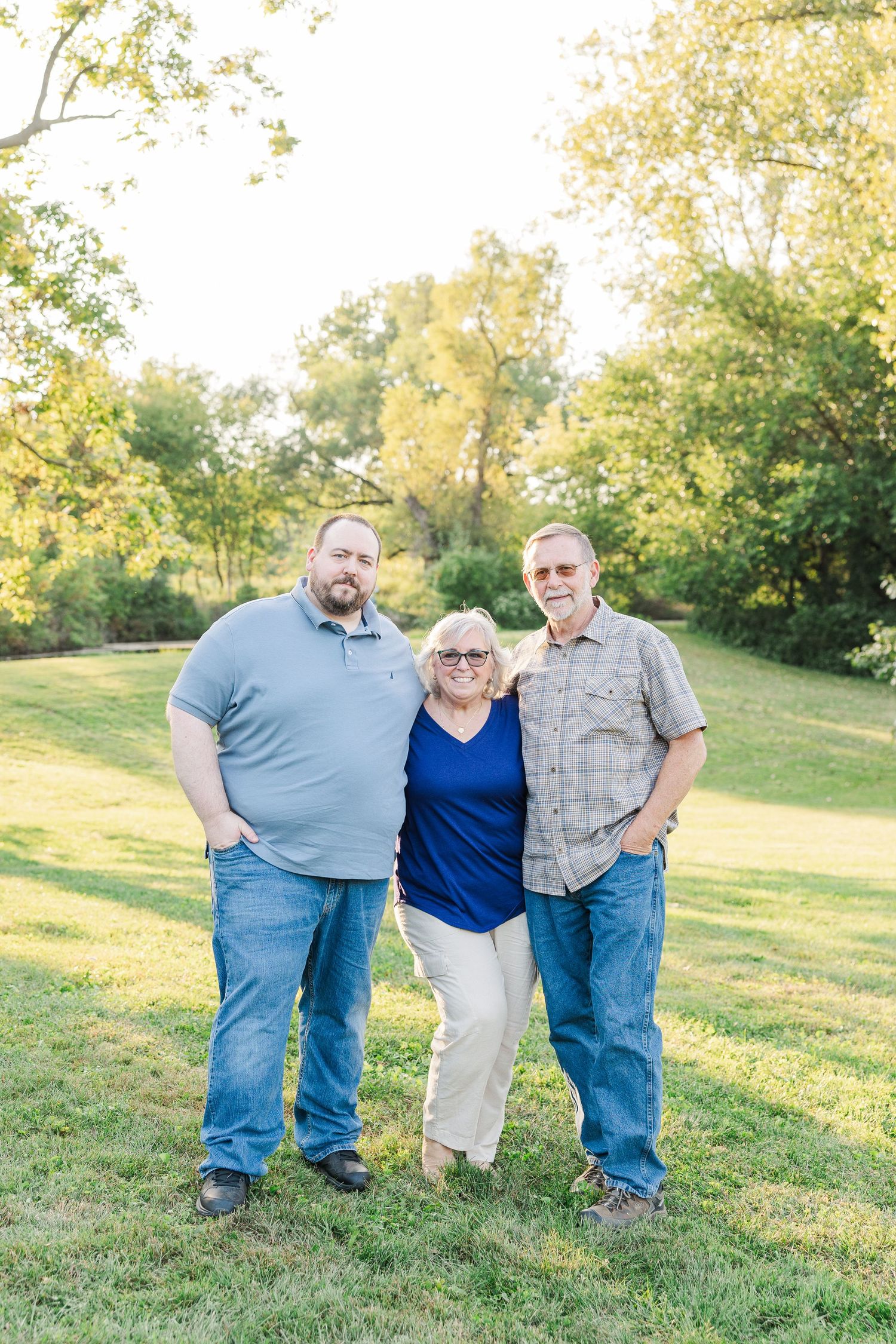 Three figures pose casually together on a grassy hillside during golden hour in a peaceful outdoor setting.Sorg family portraits at Shamrock