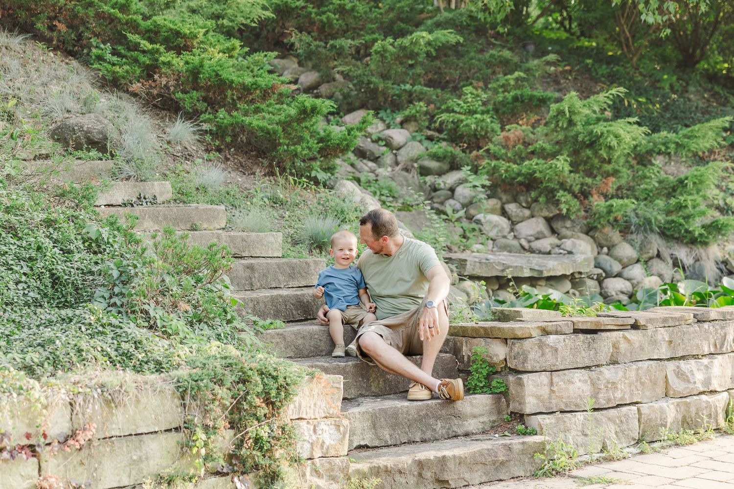 Two people walk up stone garden steps surrounded by lush greenery and landscaping on a sunny day.