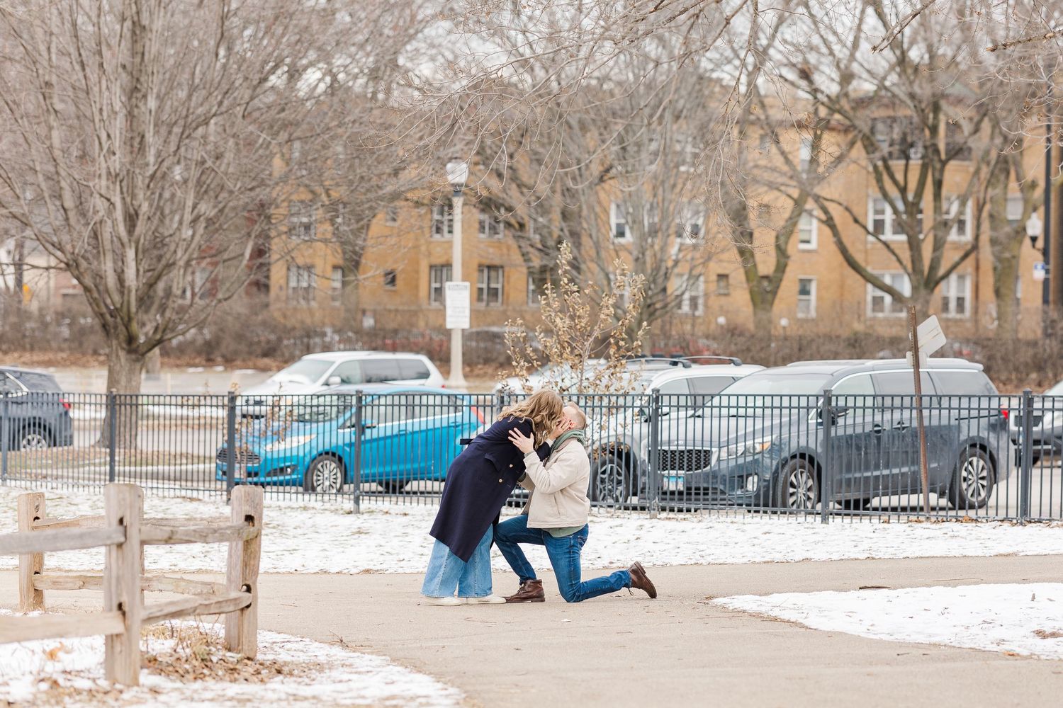 Man proposes to woman in snow-dusted park with bare trees and parked cars visible behind an iron fence in winter. Winnemac Park, Chicago