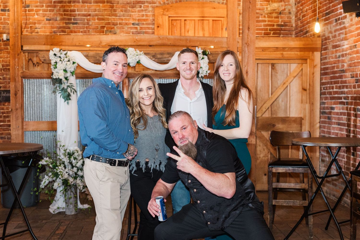 Group photo of wedding guests posing playfully in front of rustic wooden doors and brick walls.