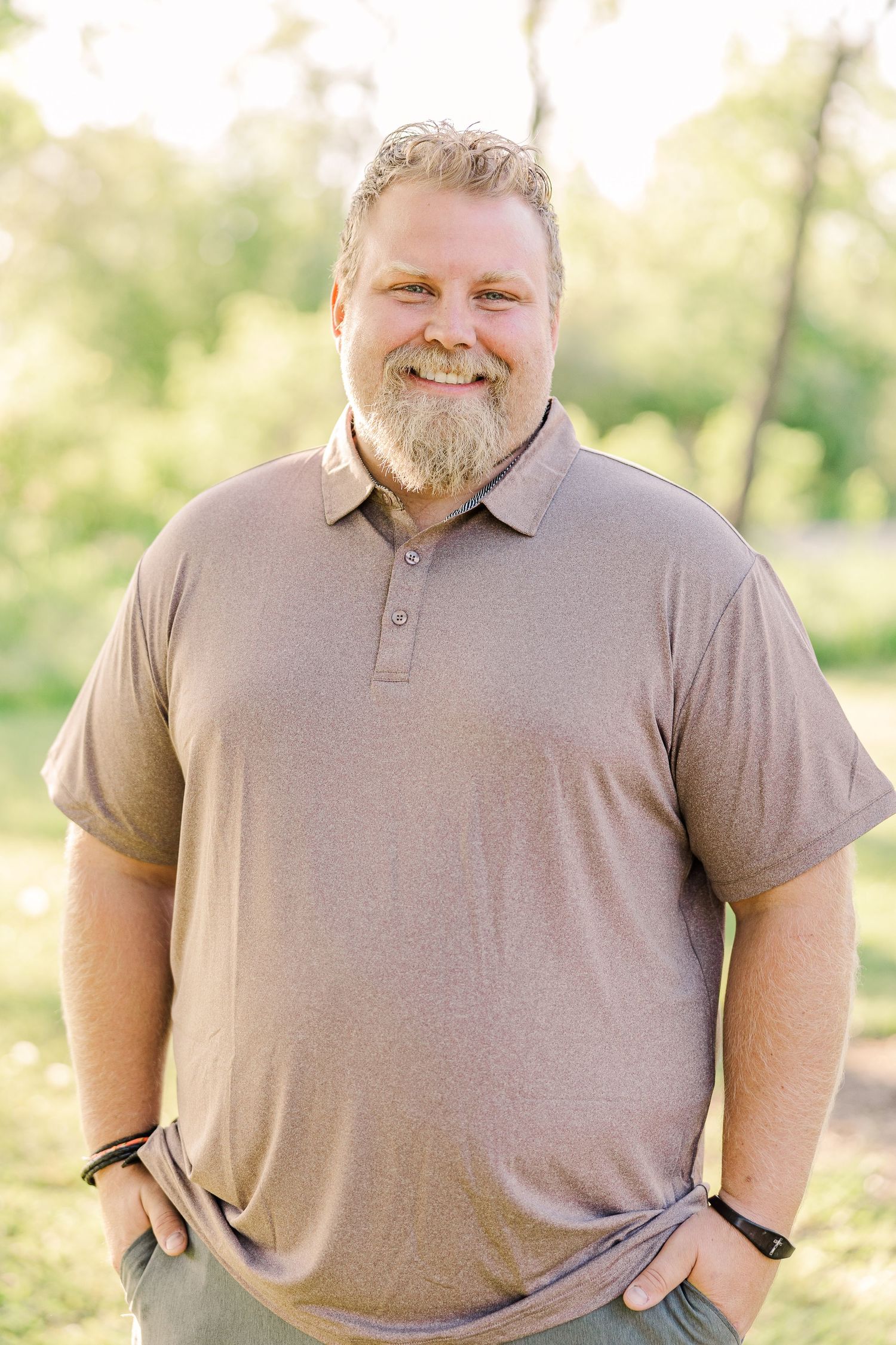 A person in a tan polo shirt smiling warmly at the camera outdoors.