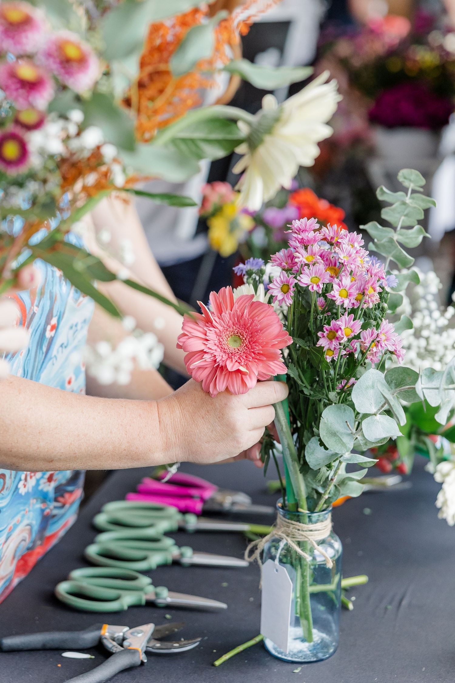 Florist arranging a pink gerbera daisy bouquet in a mason jar with eucalyptus at a flower arranging workshop.