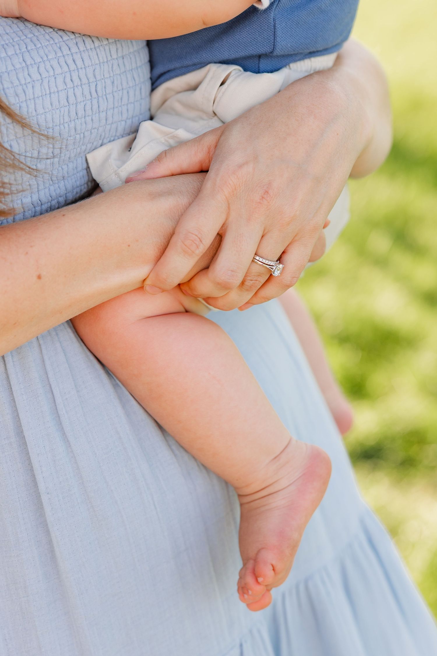 Close-up of a mother in a blue dress holding her baby, wearing a diamond engagement ring outdoors.