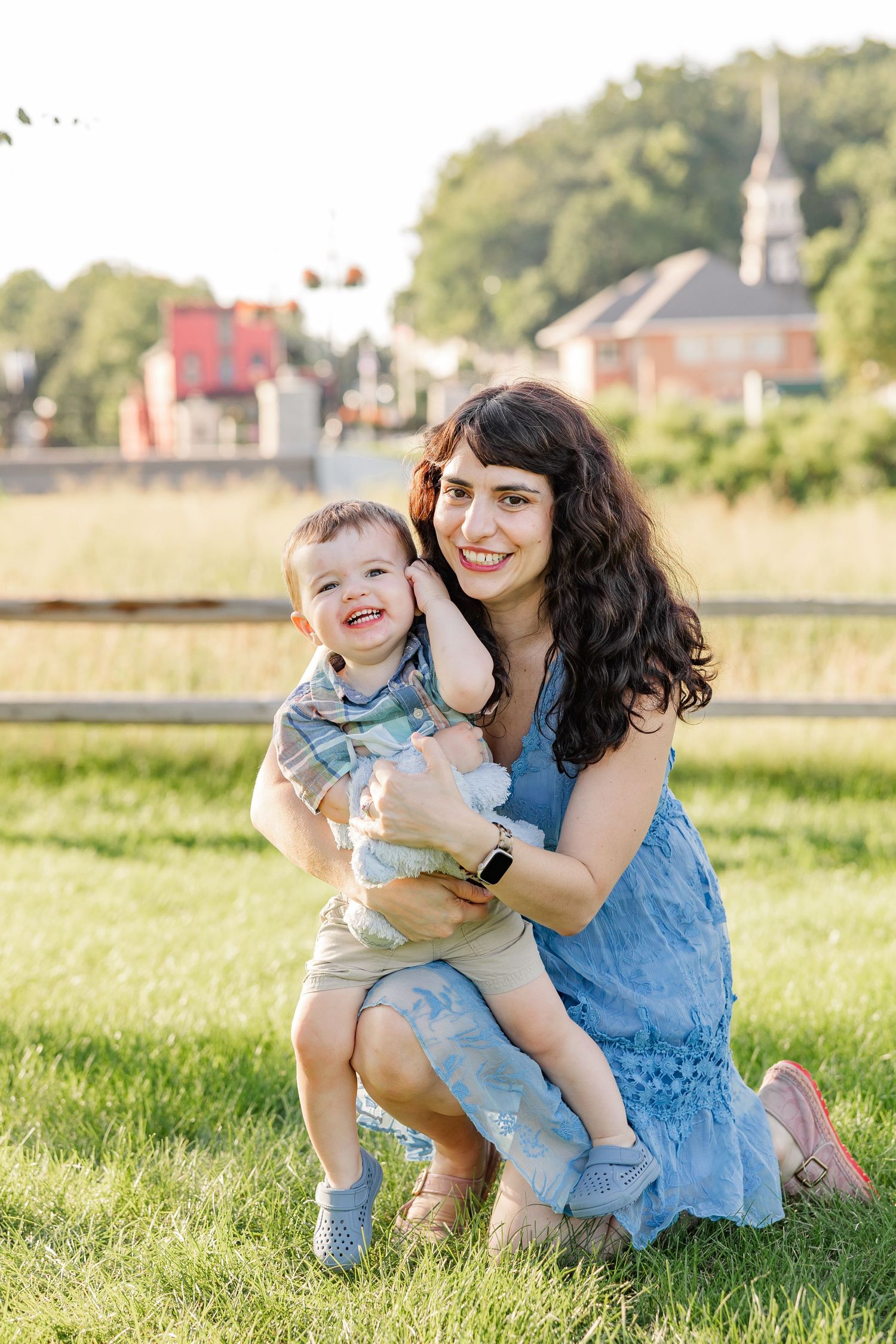 A dark-haired mother in a blue dress smiles warmly while holding her laughing toddler son outdoors in sunlight.