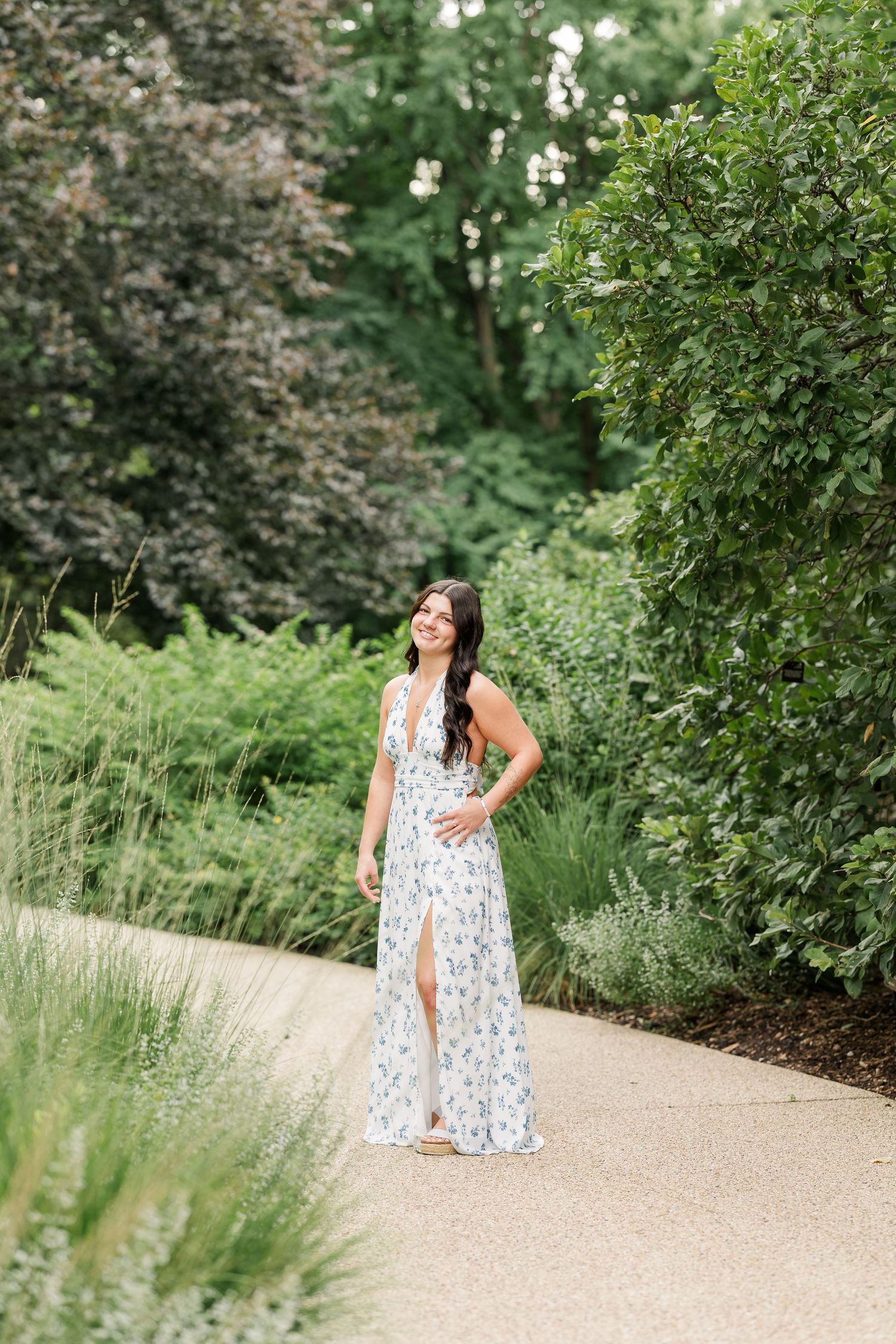 Young woman in white floral maxi dress smiling on garden path surrounded by lush greenery.