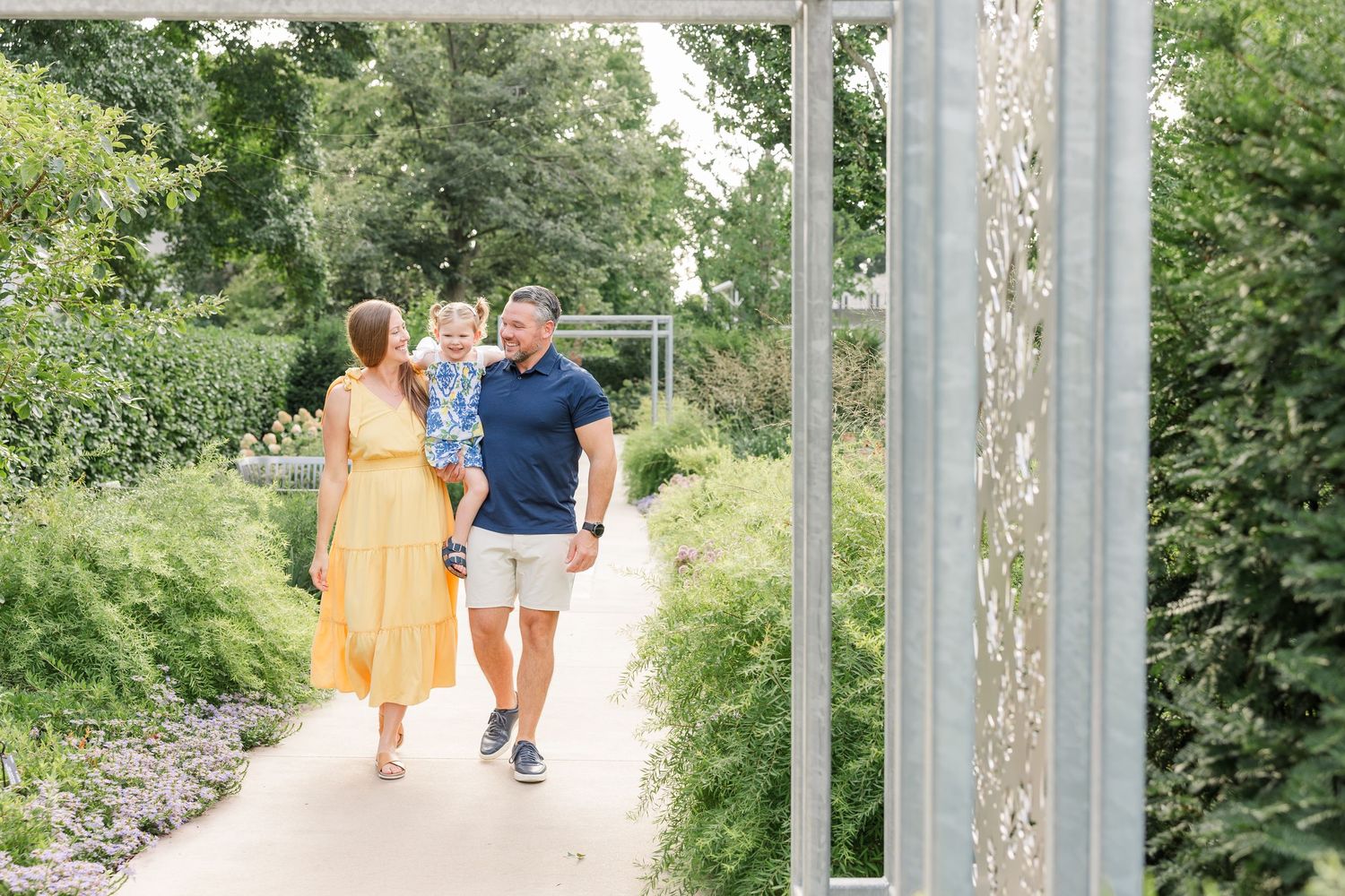 Family of three walking along a garden path framed by lush greenery.