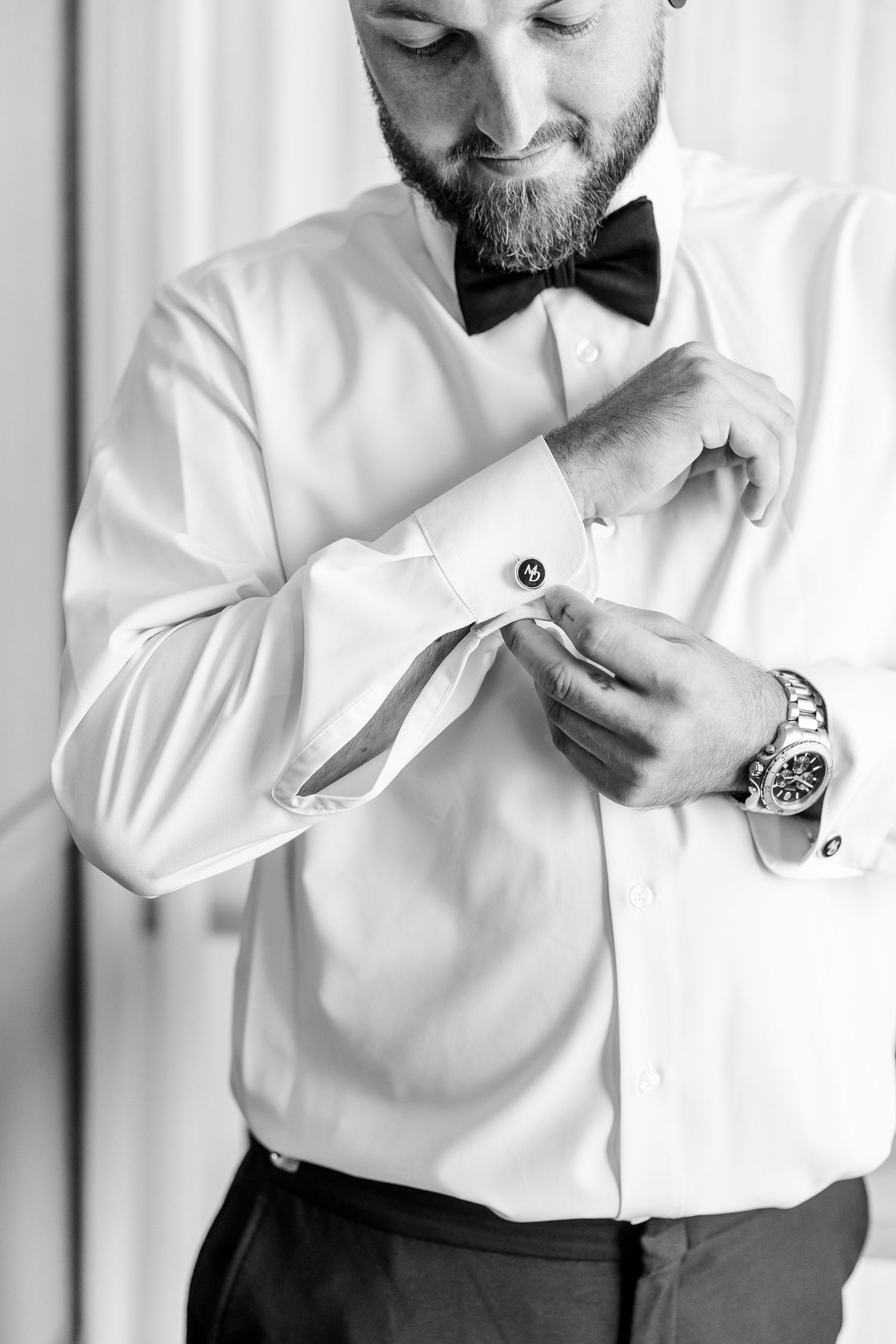 Black and white portrait of bearded groom in white shirt fastening cufflinks before wedding.