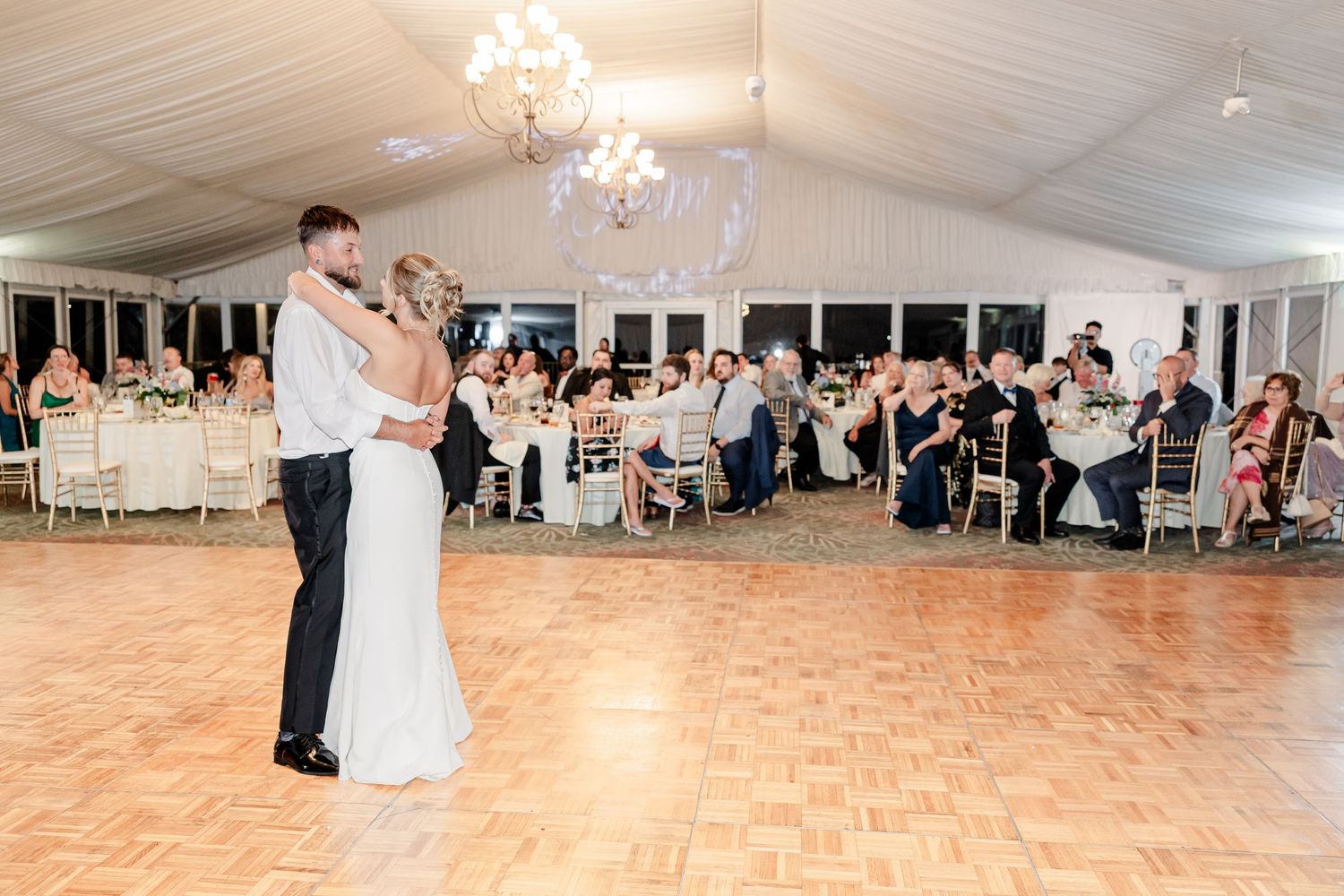 Bride and groom share first dance in elegant white tent reception hall surrounded by seated wedding guests.