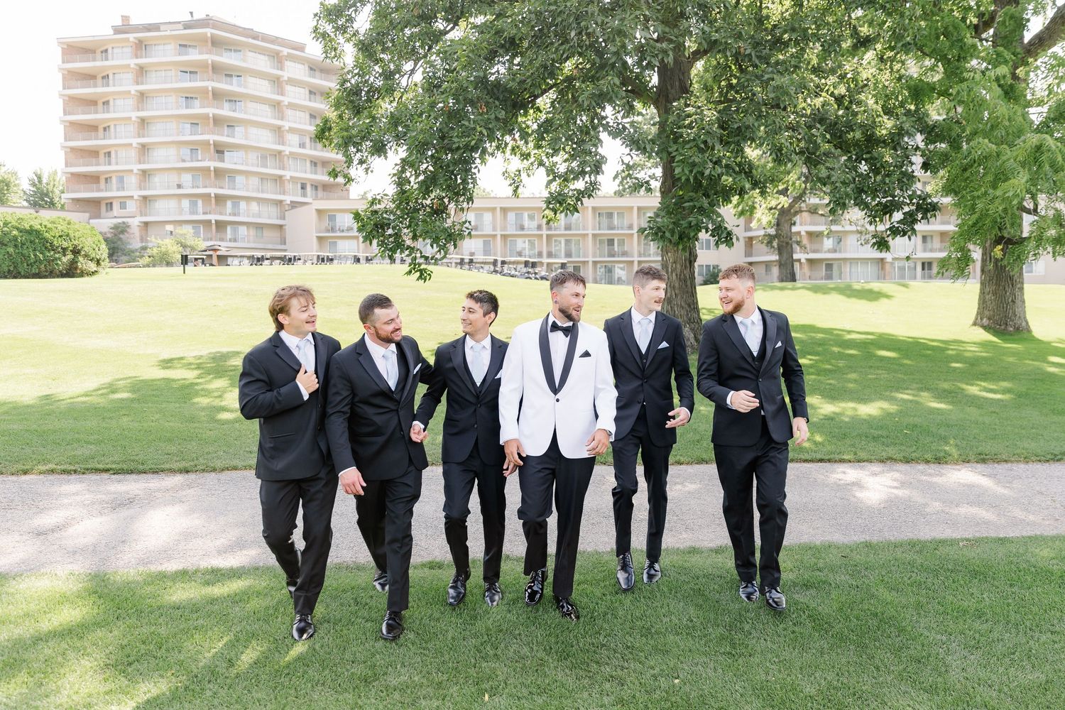 Five groomsmen in black tuxedos walk together outdoors on a sunny day, with the groom in a white jacket at center.