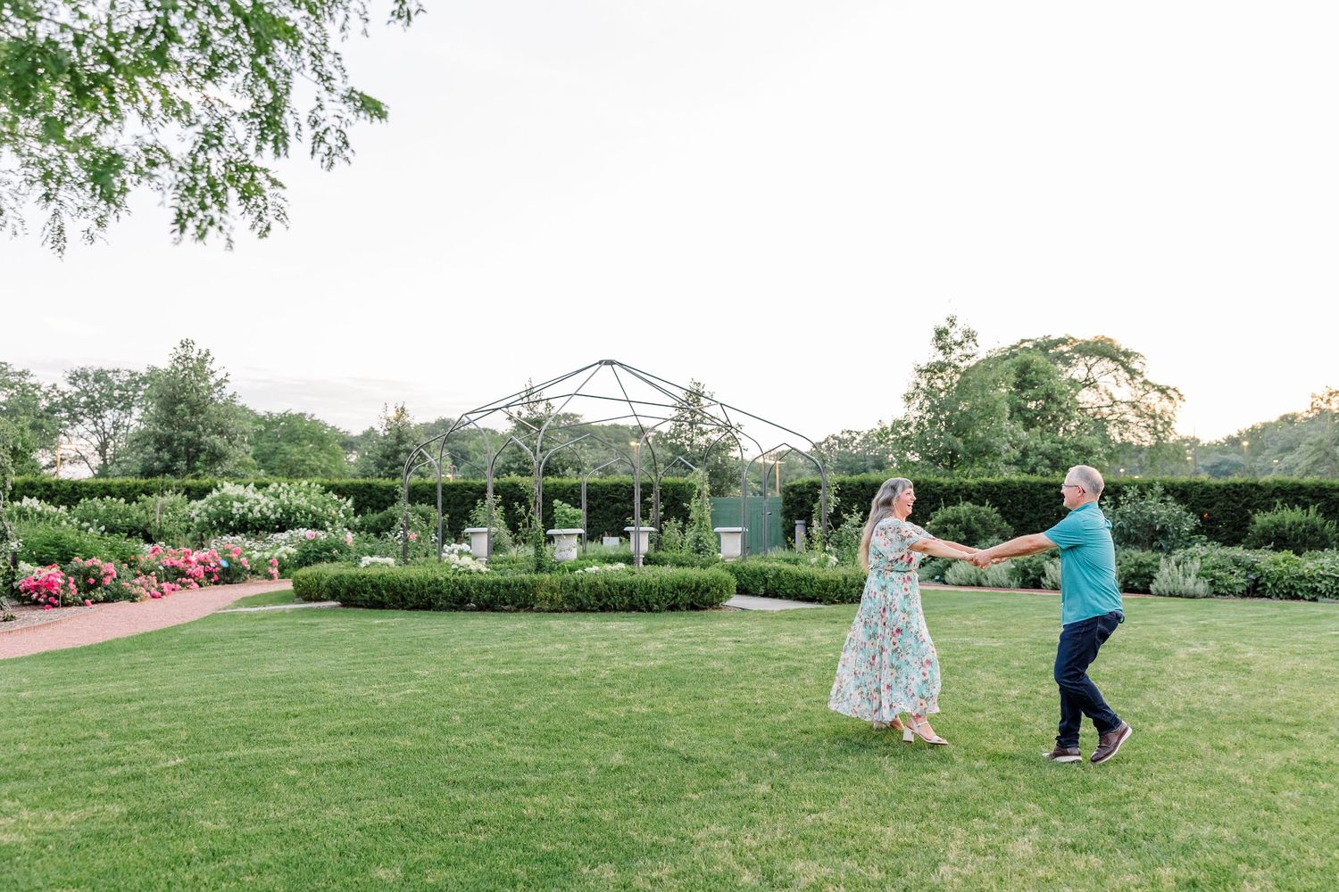 Couple holding hands and dancing on a wide green lawn with elegant garden greenhouse in the background.