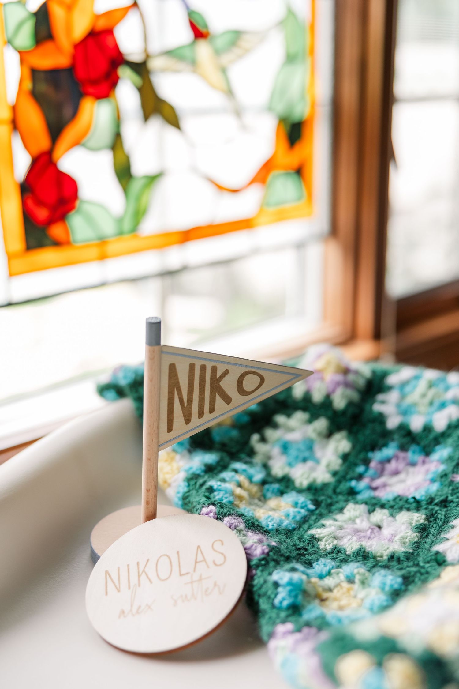 Wooden pennant flag and round sign reading Nikolas beside colorful crocheted blanket near stained glass window.