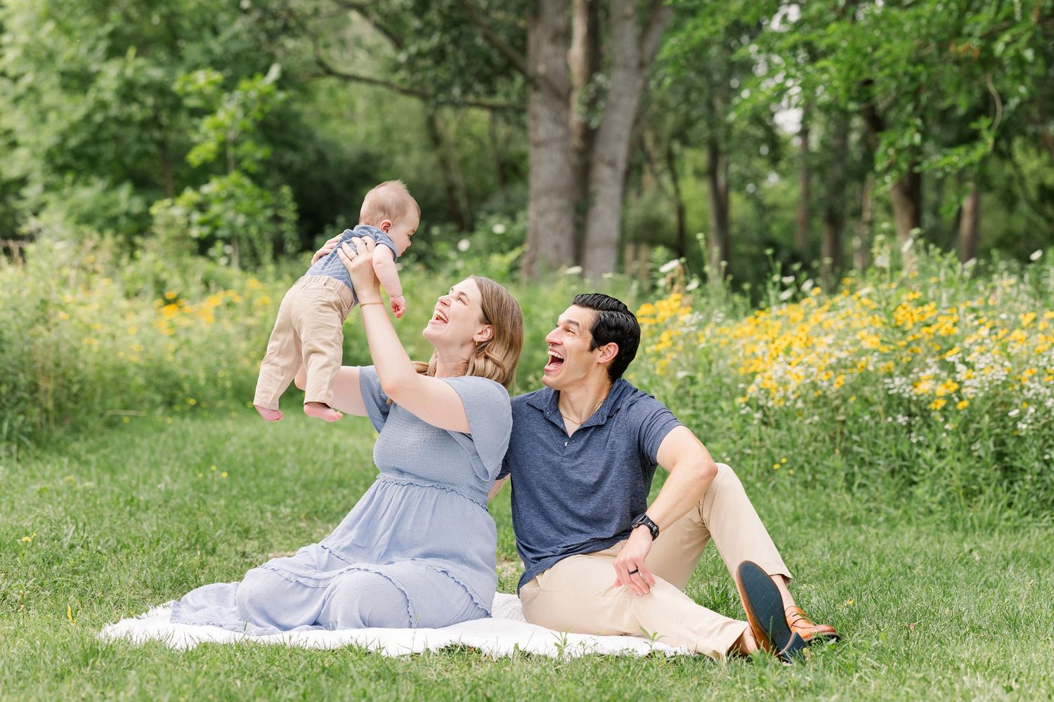 Young couple sits on grass laughing while lifting baby in sunny park with wildflowers.