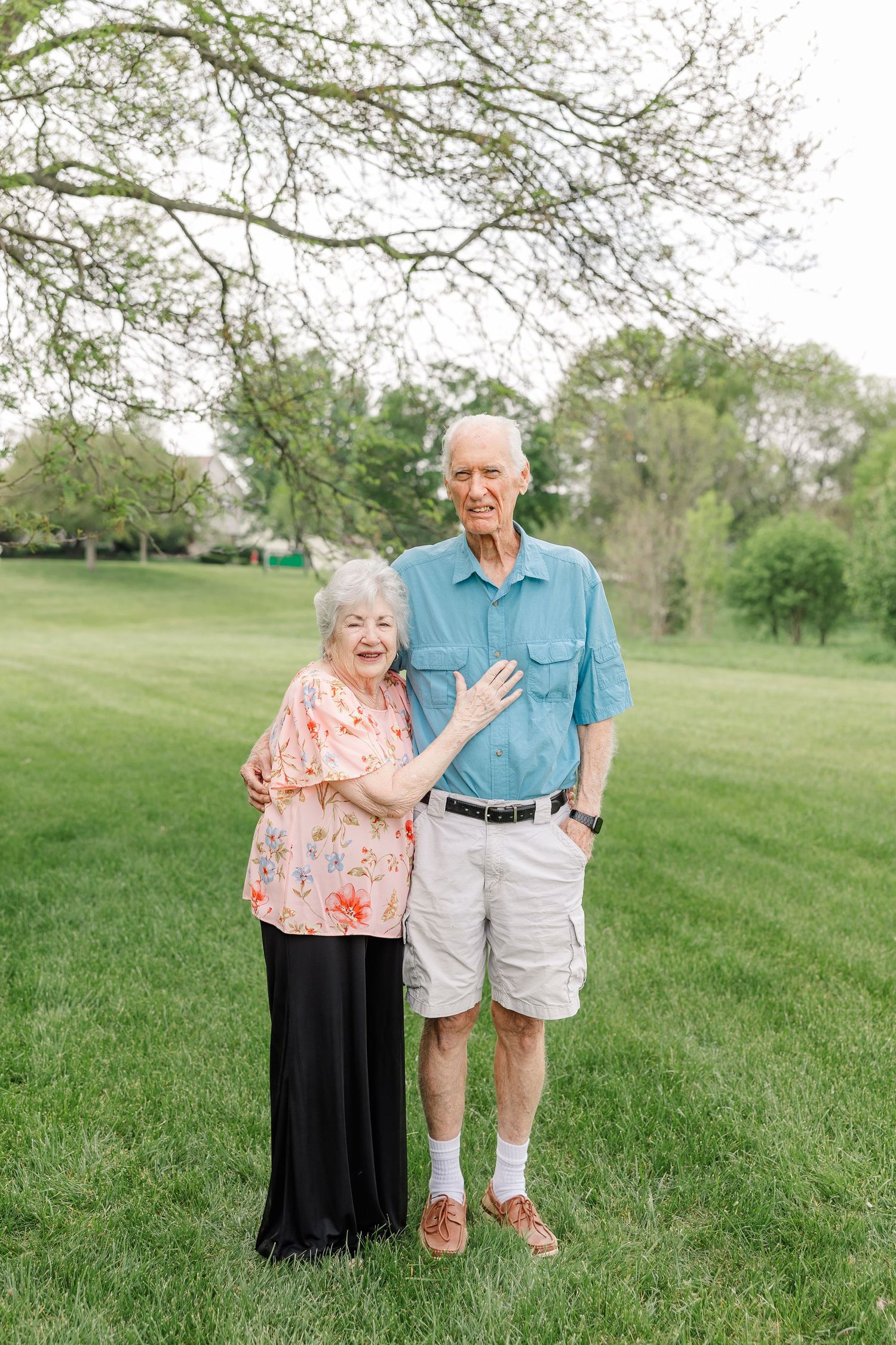 Happy elderly couple standing together on green lawn under bare tree branches during outdoor portrait session.