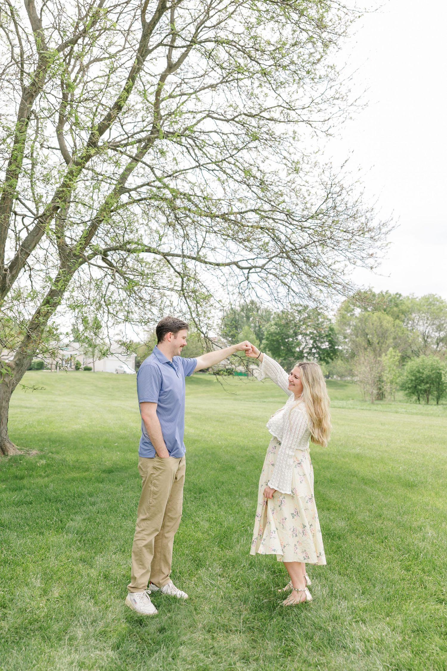 Young couple dancing outdoors near blooming tree, man spinning woman in floral skirt on green lawn.