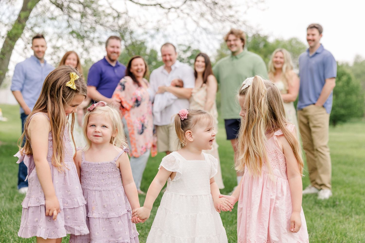 Young girls in pastel dresses hold hands outdoors while family members gather and smile in a lush green park setting.