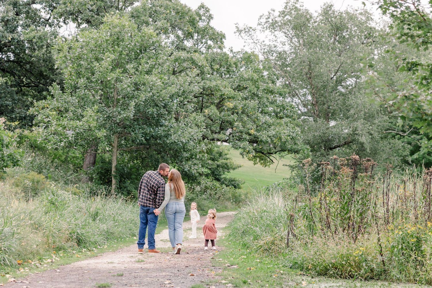Family with toddler walking on a dirt path surrounded by lush green trees and meadows on a cloudy day.