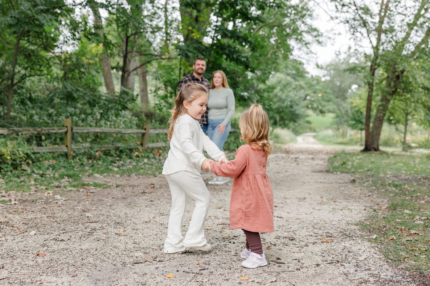 Two young girls hold hands and spin on a dirt path while their parents watch lovingly in a lush green outdoor setting.