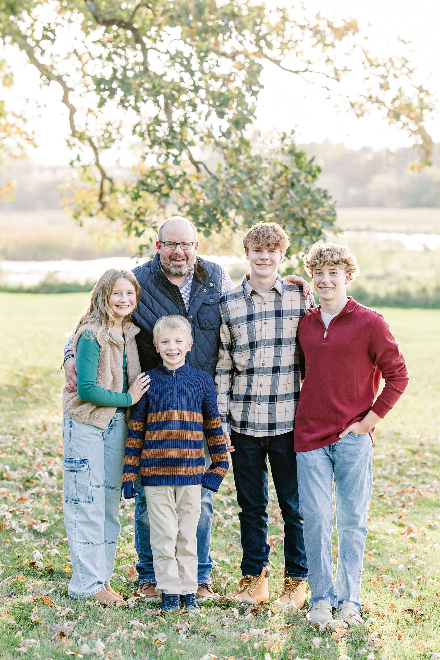 A smiling family of five stands together on an autumn lawn under a tree, with a bearded dad, a girl, and three boys posing.