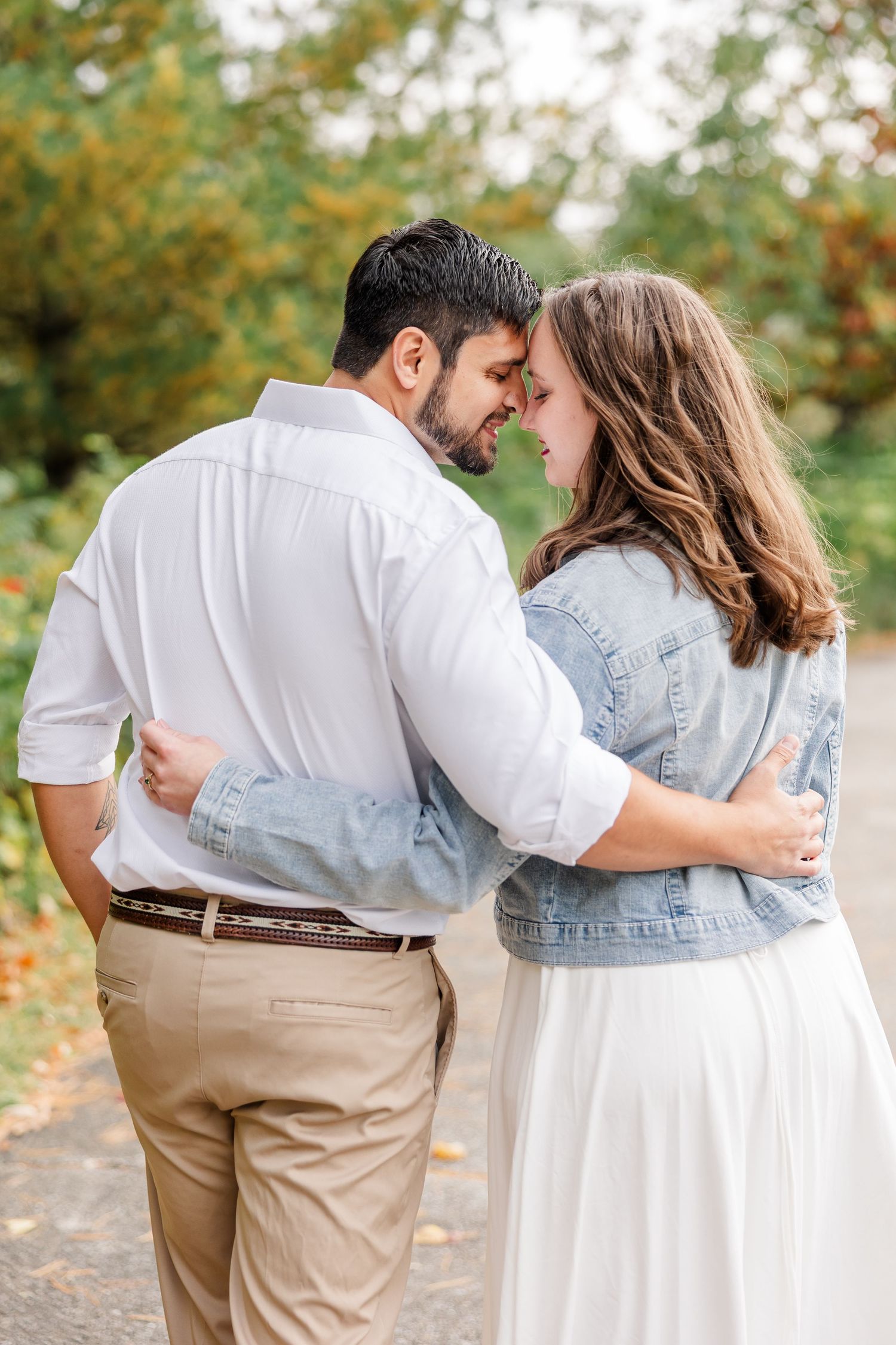 Couple in white shirt and denim jacket embracing with foreheads touching on a leaf-lined path during an autumn engagement session.