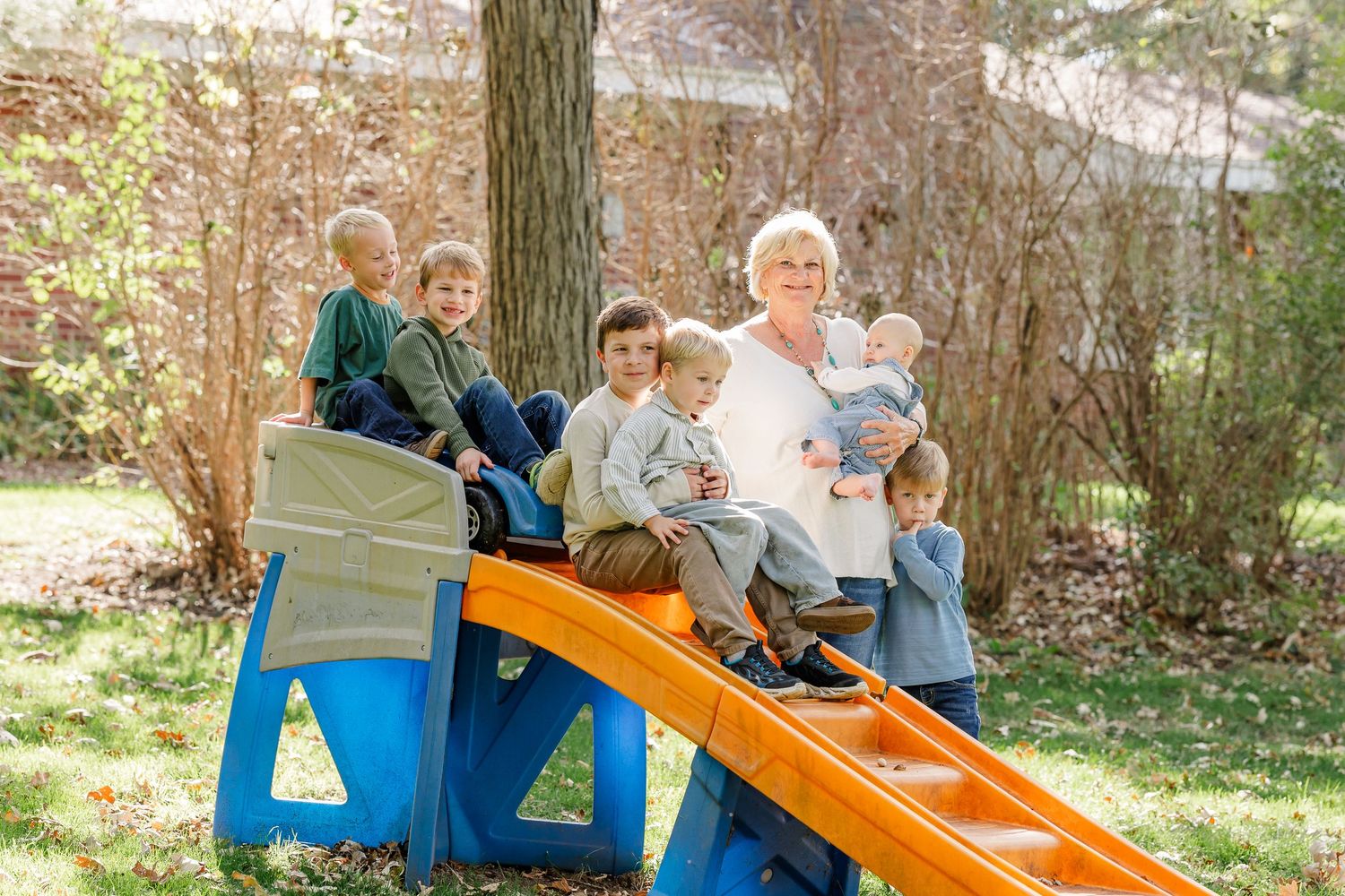 Grandmother sits on a colorful slide surrounded by five young grandchildren outdoors.