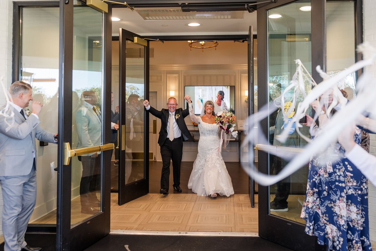 Newlywed bride and groom raise their arms in celebration as they walk through open glass doors while guests cheer and toss streamers.