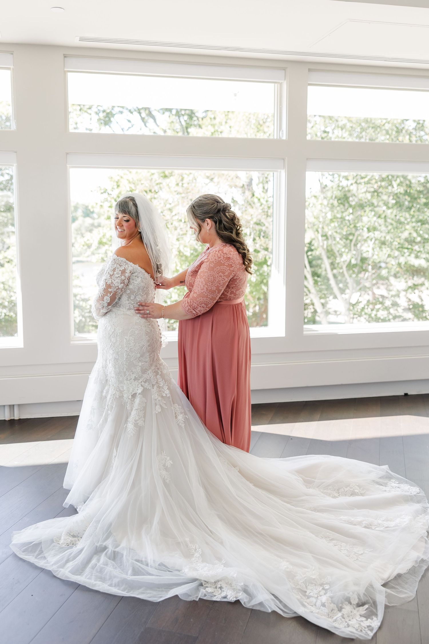 Bridesmaid in a dusty rose dress helps button the lace wedding gown of a bride with a long train in a bright room with large windows.