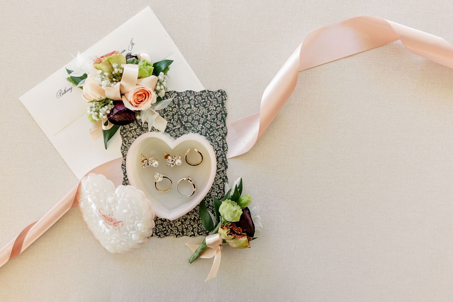Wedding rings in a heart-shaped dish with peach flowers, a boutonniere, and a blush ribbon on a textured invitation flat lay.