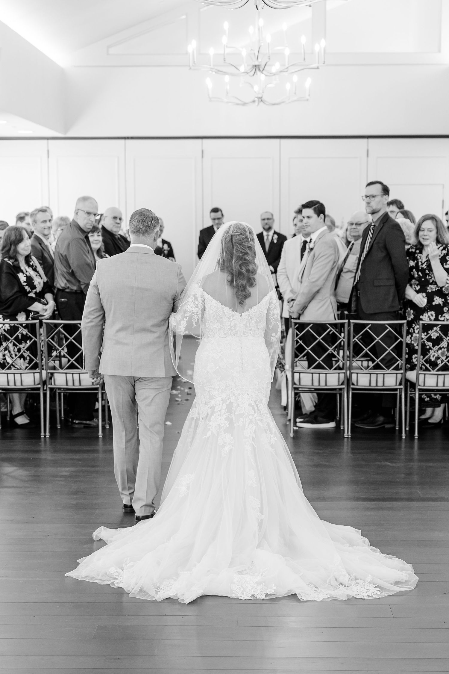 Black and white photo of a bride in a lace gown with a long train walking down the aisle with her father toward seated wedding guests.